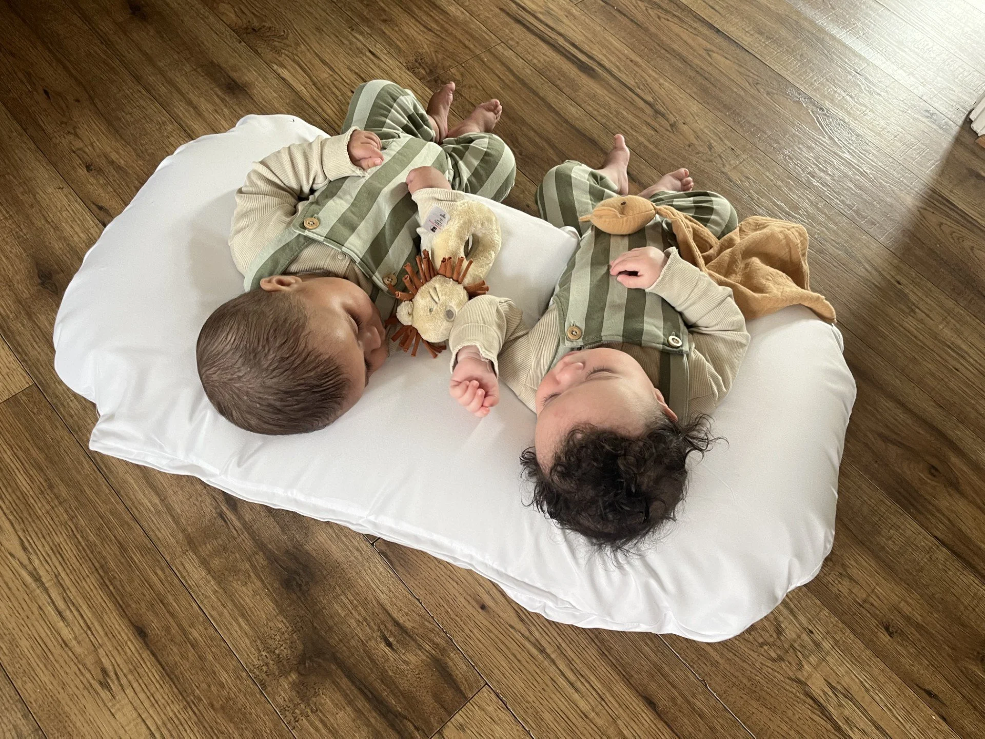 Twin babies lying side by side on a white support pillow during supervised playtime.
