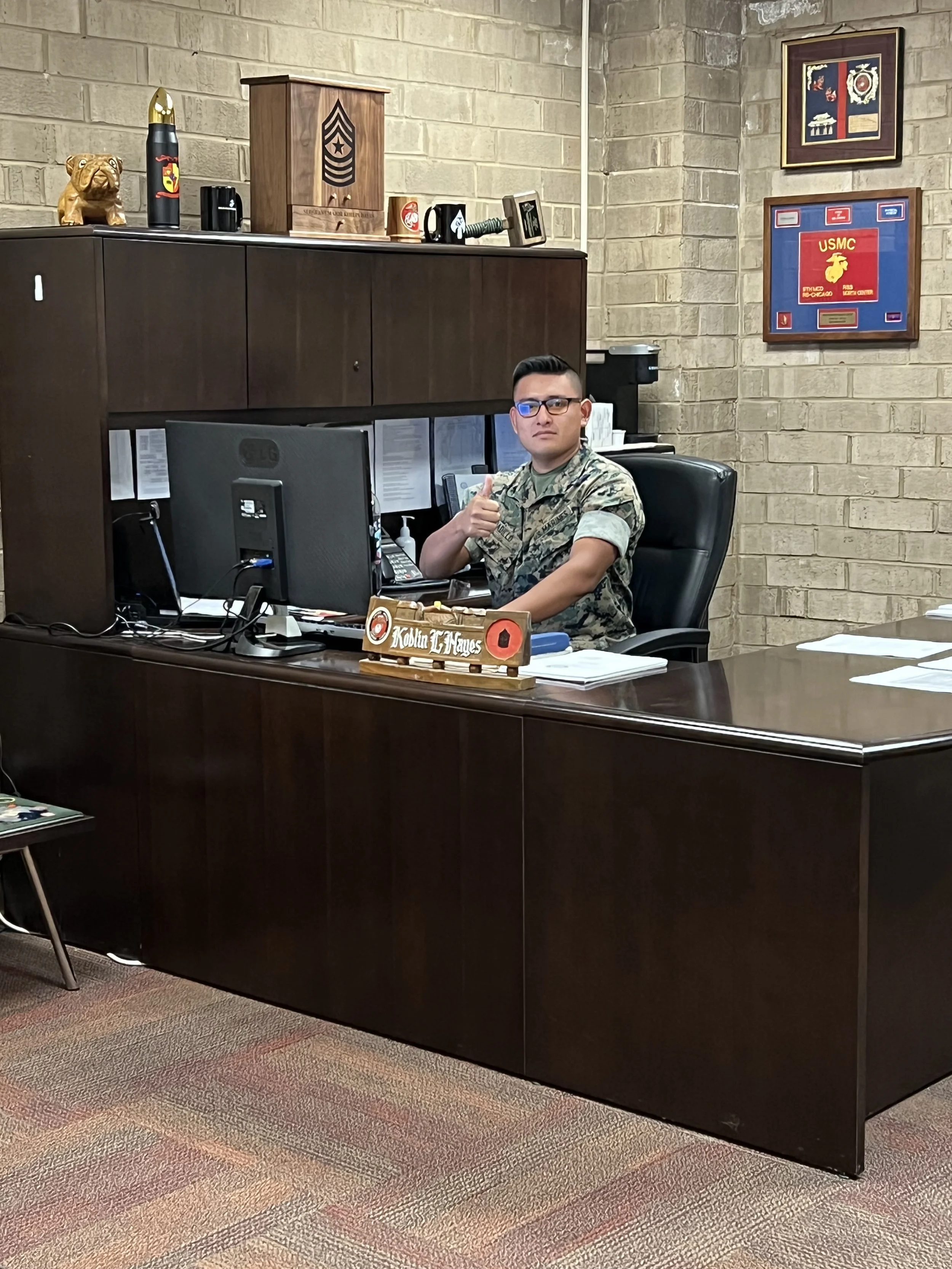 A man in military camouflage uniform sitting at a dark brown office desk with a computer, giving a thumbs-up gesture, in a room with beige brick walls decorated with military memorabilia and framed pictures.