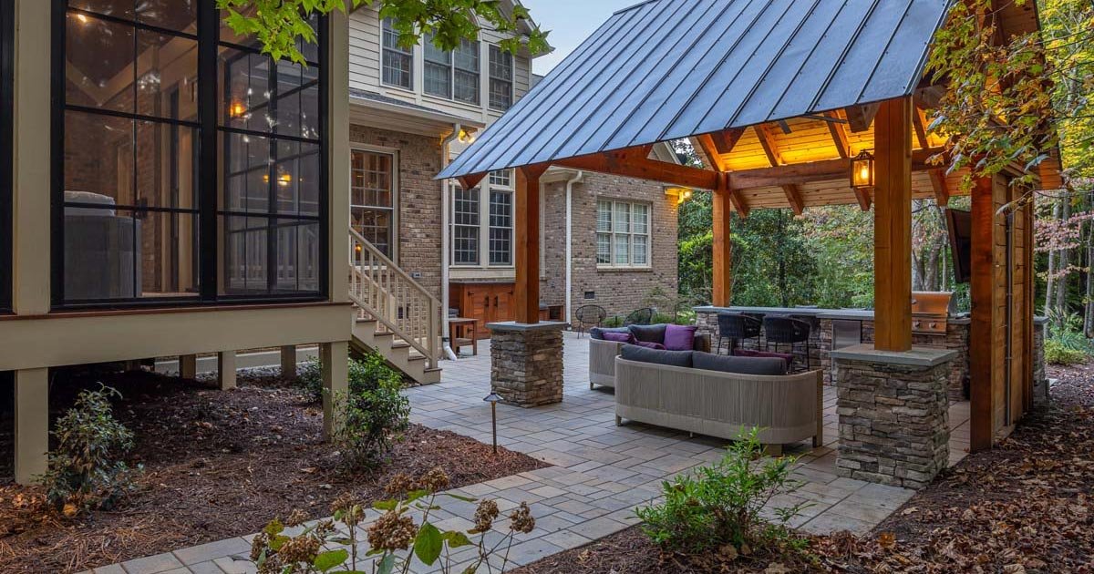 A backyard patio area with a covered wooden structure, outdoor sofa, chairs, and bar seating, surrounded by trees and garden beds, attached to a brick house.