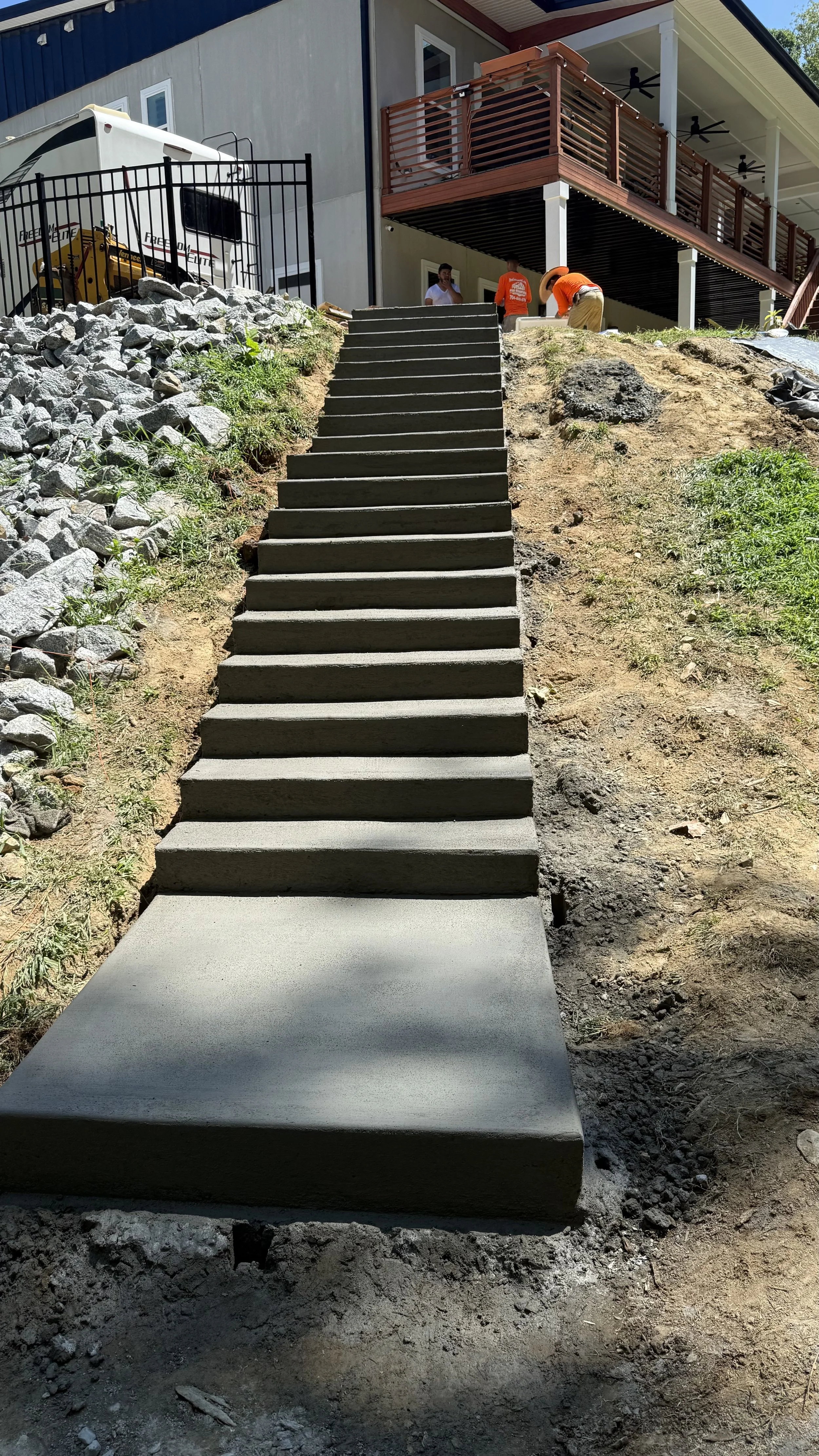 Concrete stairs leading up a slope to a wooden deck of a house with people working on construction.