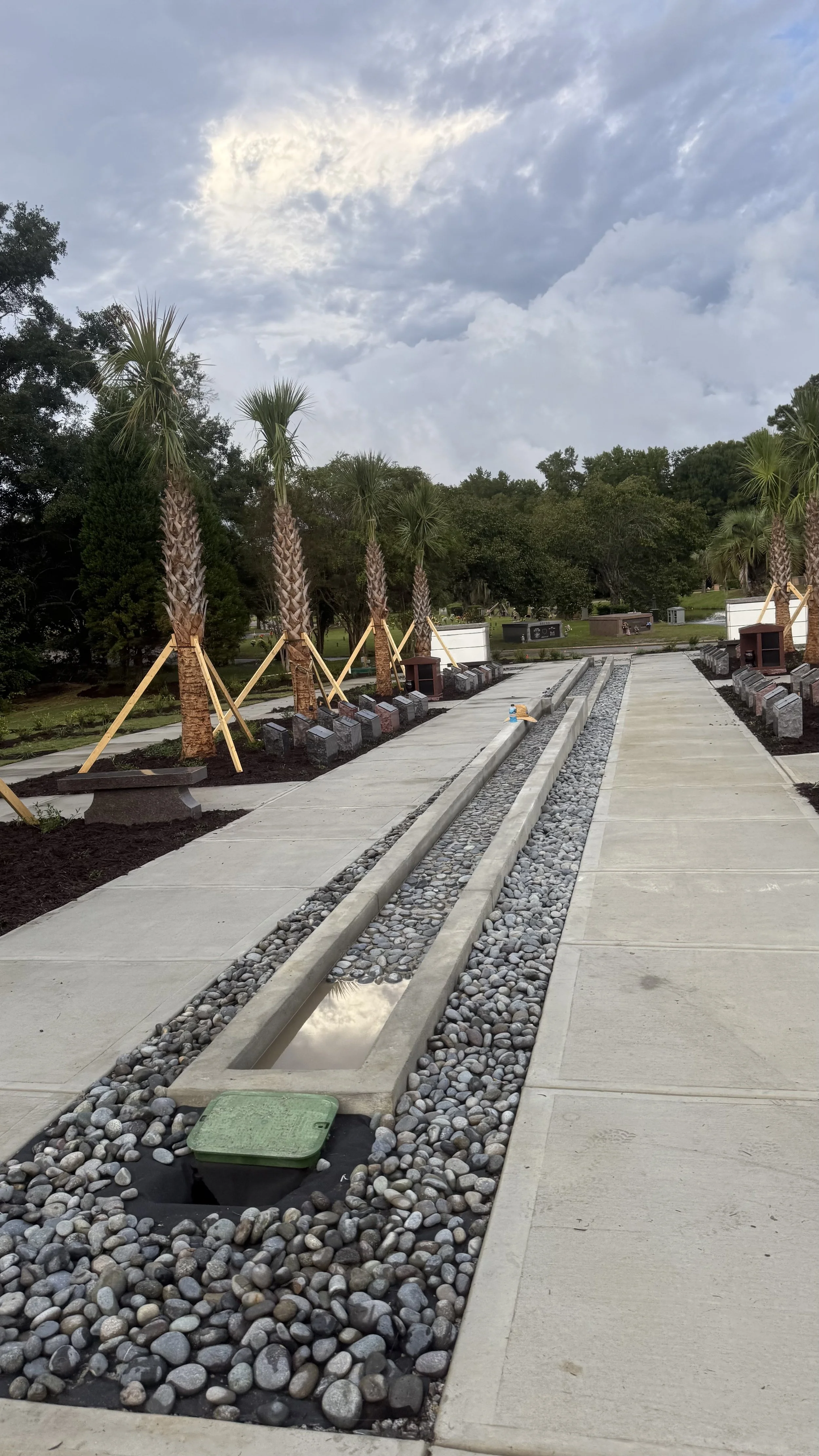 A pathway under construction with a narrow water feature filled with smooth gray stones, lined with young palm trees on one side and extending into a park with trees and benches in the background. The sky is cloudy with patches of sunlight.