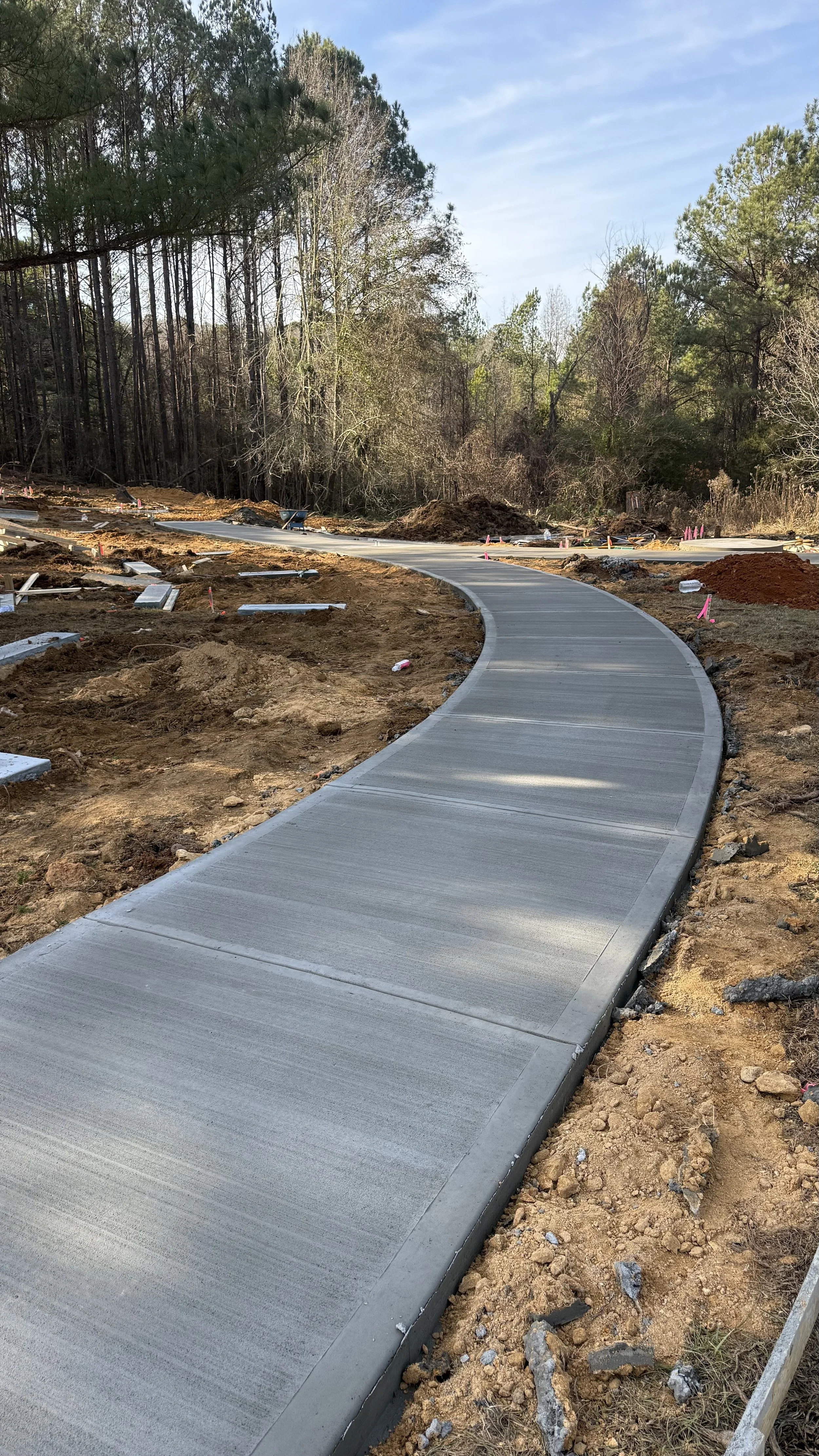 Freshly poured concrete sidewalk under construction in a natural setting with trees in the background and construction materials scattered around.