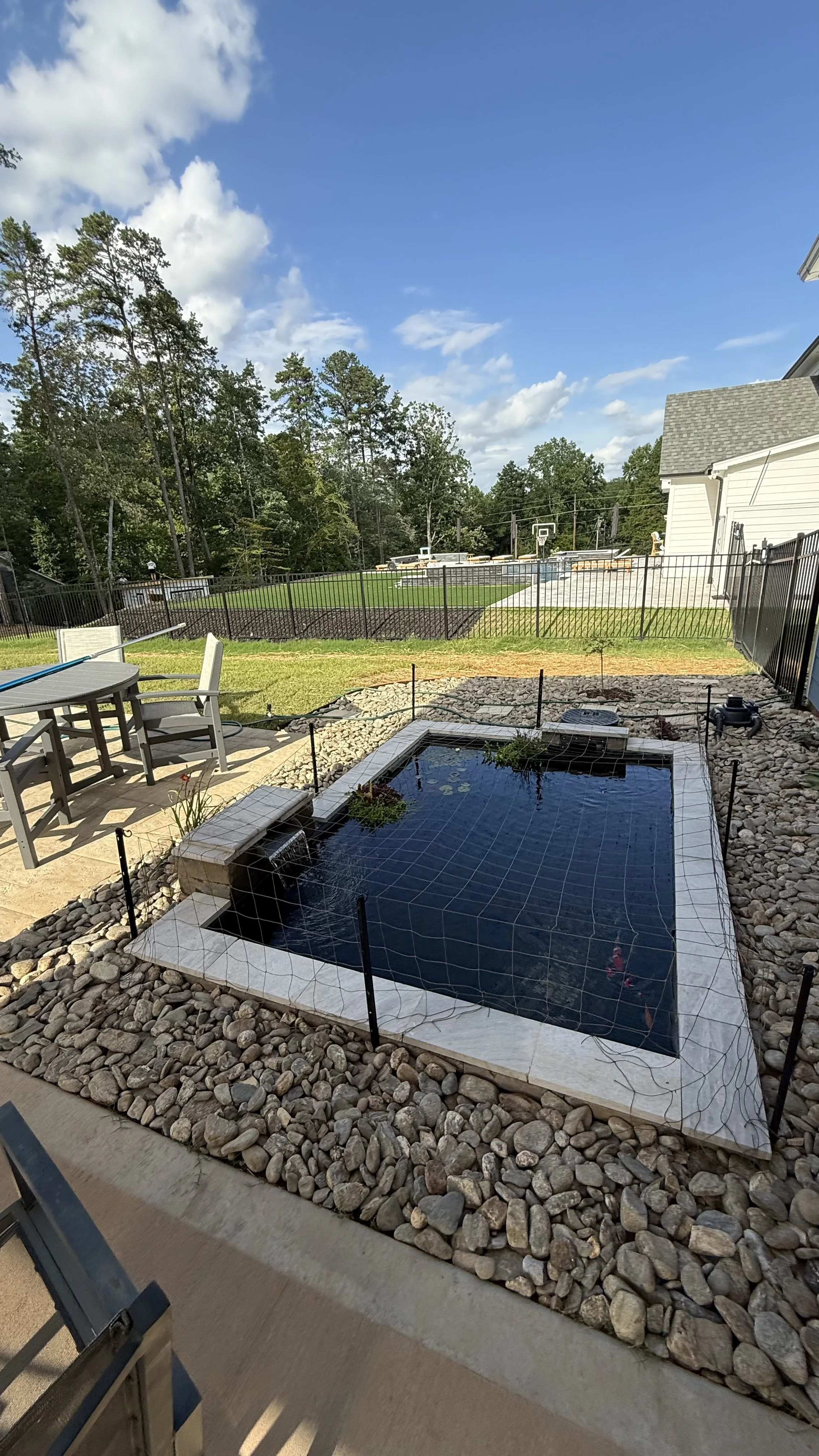 Backyard with a small pond or water feature surrounded by rocks, outdoor furniture, and a fenced sports court with a basketball hoop in the distance, under a blue sky with clouds.