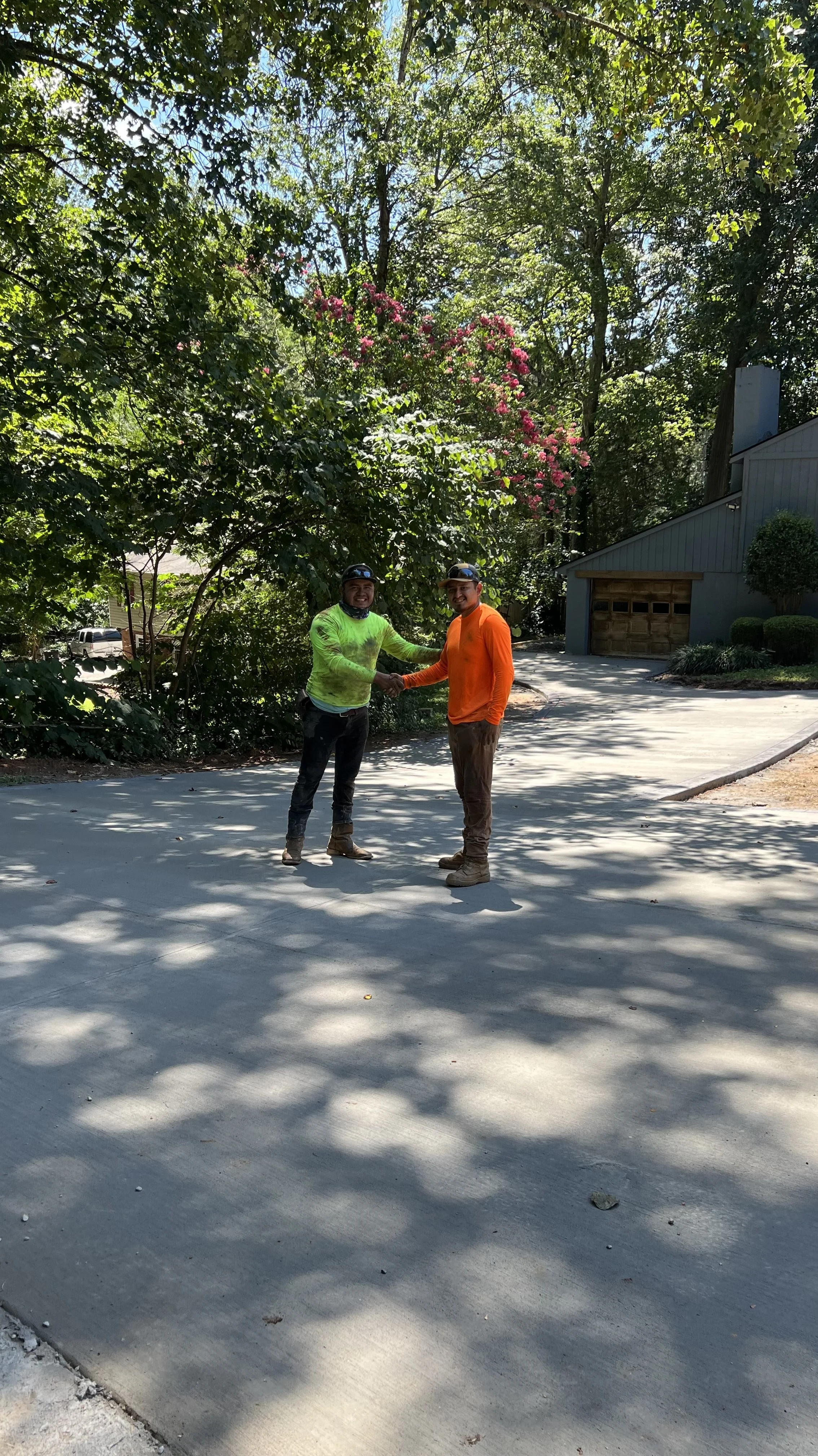 Two men shaking hands outdoors on a concrete driveway, surrounded by lush green trees and a house in the background, bright sunlight casting shadows.