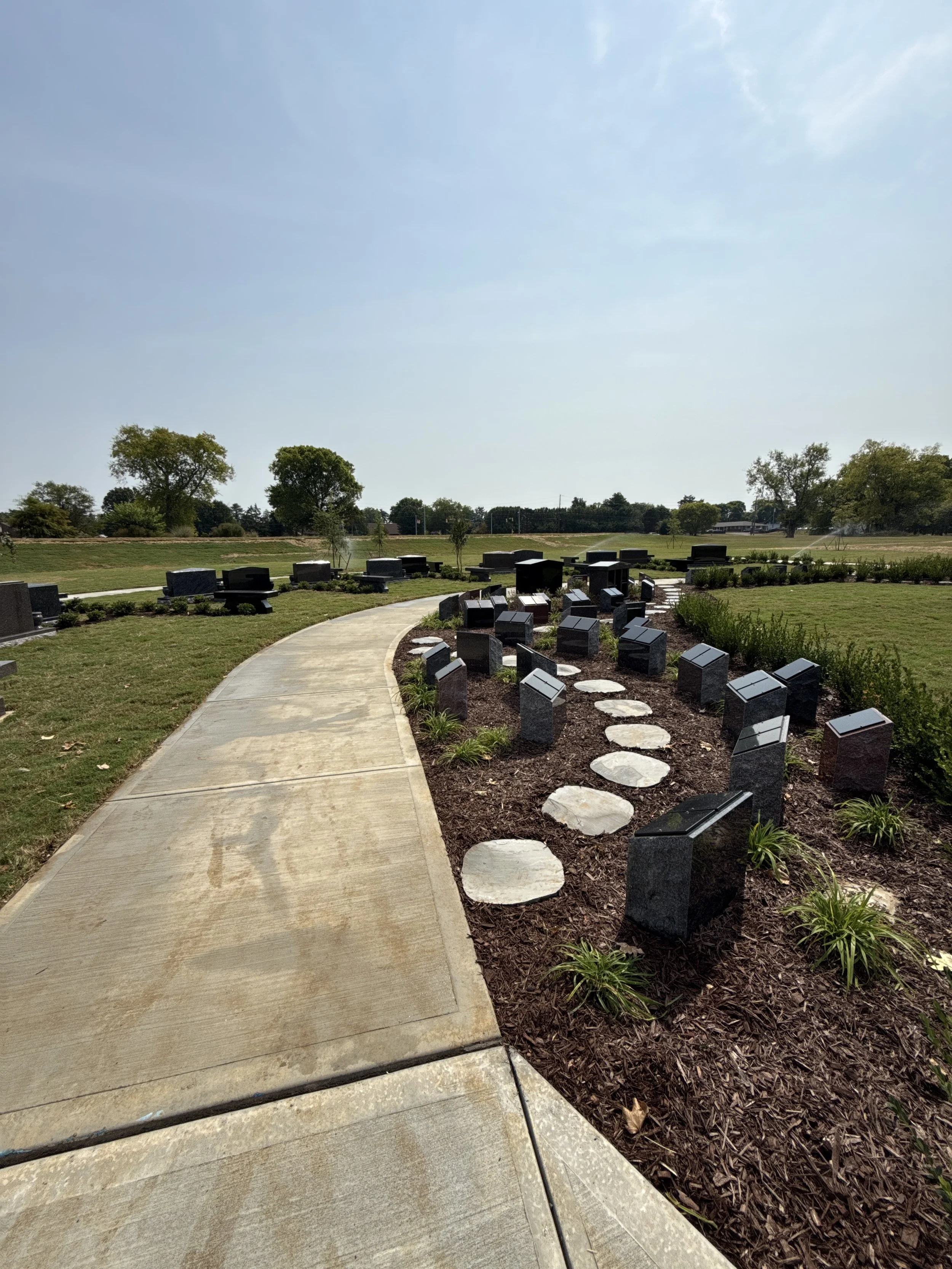 A sidewalk winding through a landscaped memorial site with black granite headstones, surrounded by green grass and trees, under a clear blue sky.