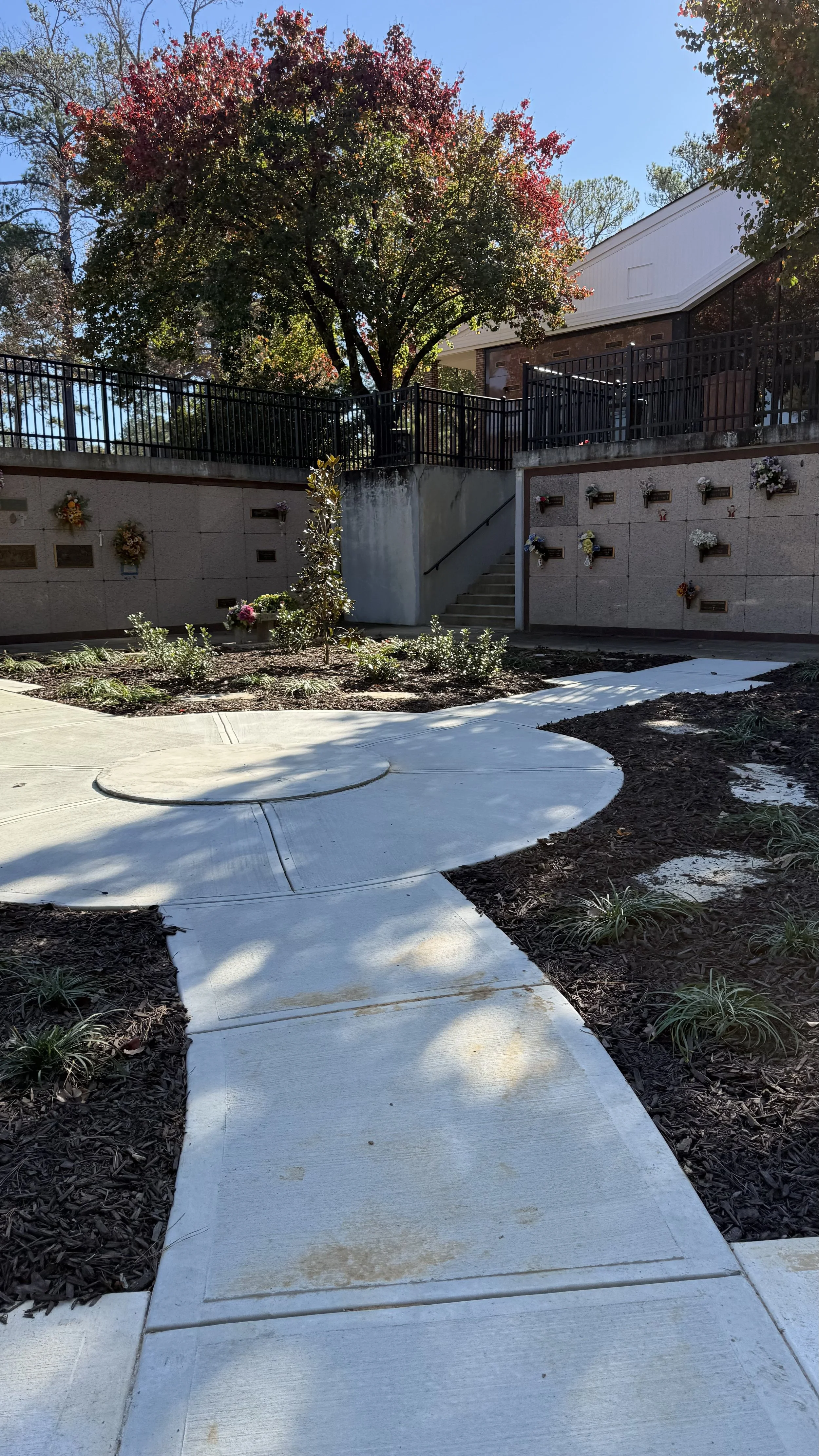 Concrete walkway with a circular design section, leading to stairs in a garden area with flowers and bushes, and a memorial wall with plaques and flowers, in front of a building with a metal railing and a large tree with red and green leaves.