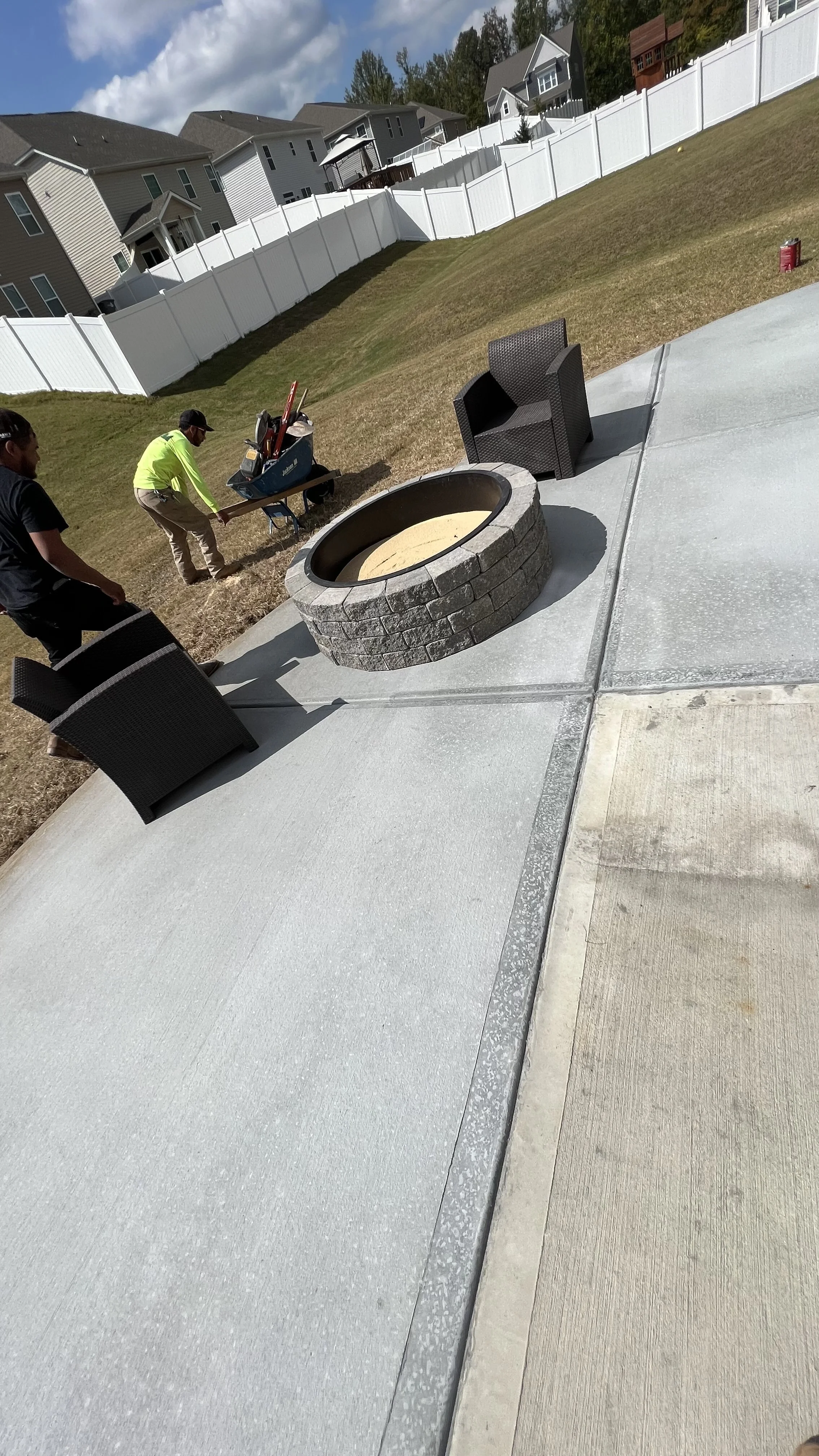 Two workers installing concrete on a backyard patio, with outdoor chairs, a fire pit, and a white fence with houses and trees in the background.