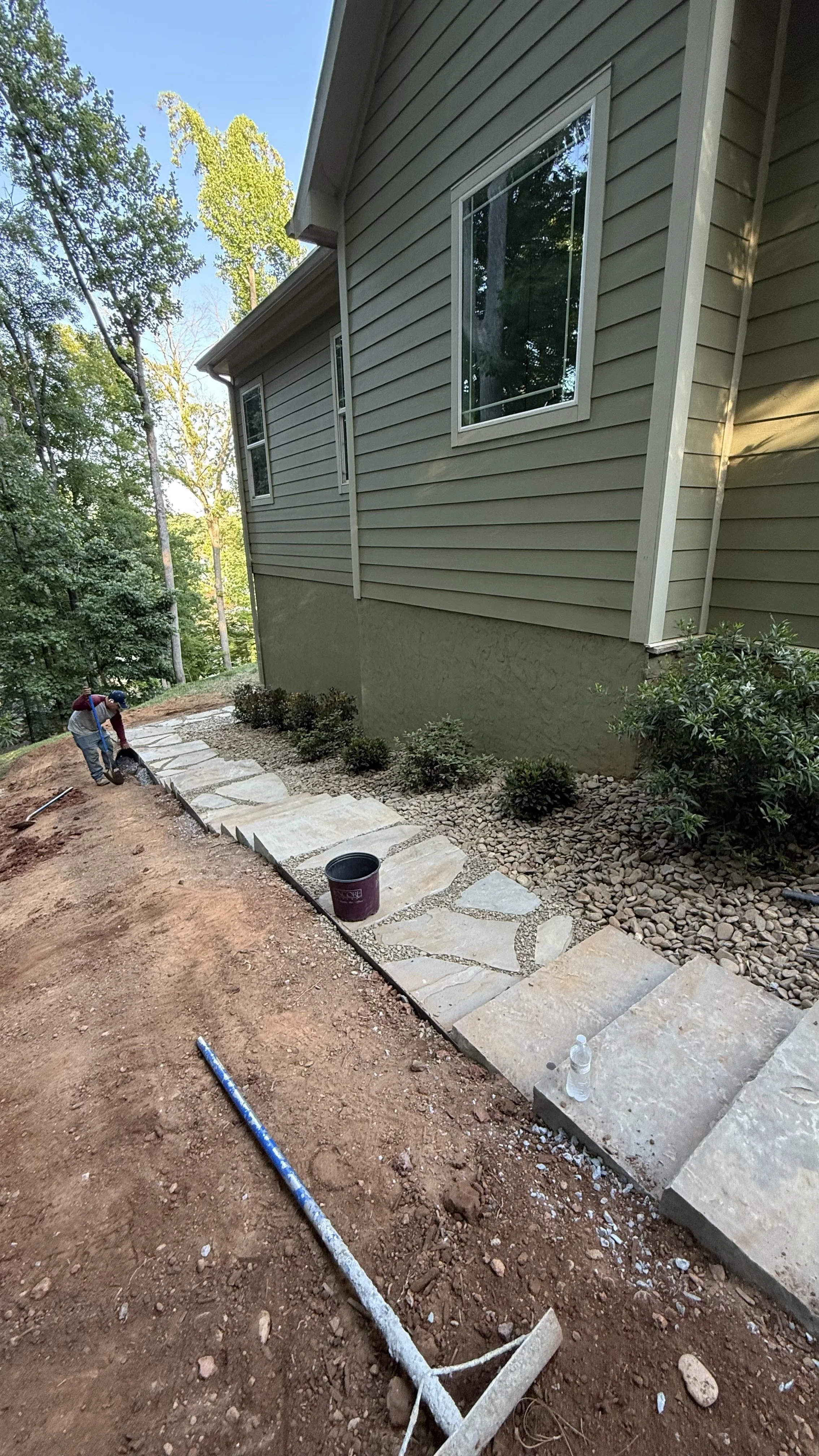 A person working on a stone pathway outside a house with beige siding and large windows, surrounded by small bushes and trees.
