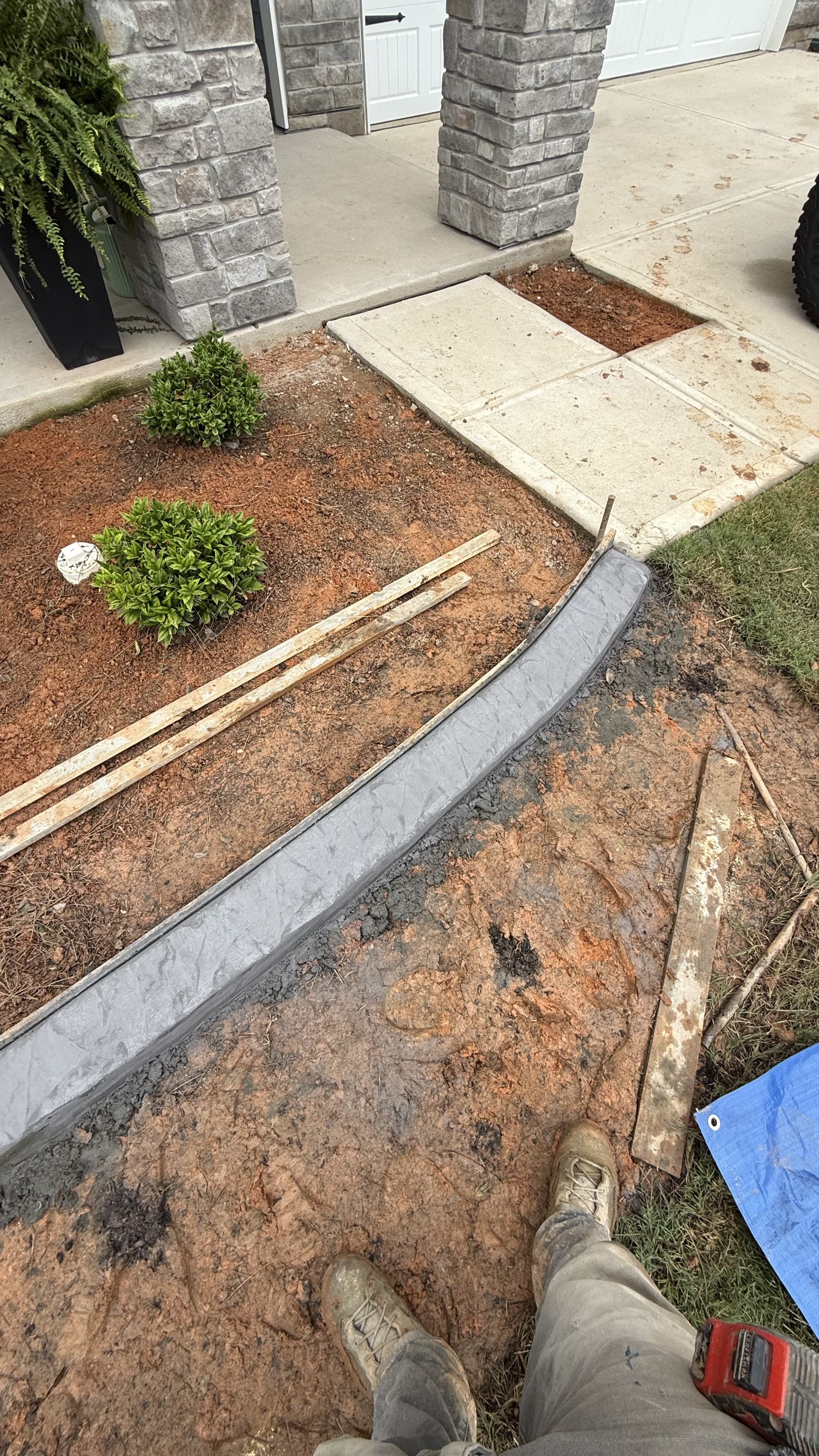A person standing on muddy ground with two plants nearby, construction work in progress for a sidewalk or pathway. A curved metallic edge is installed on the ground, with tools and wooden planks nearby, near a house with stone pillars and a garage door.