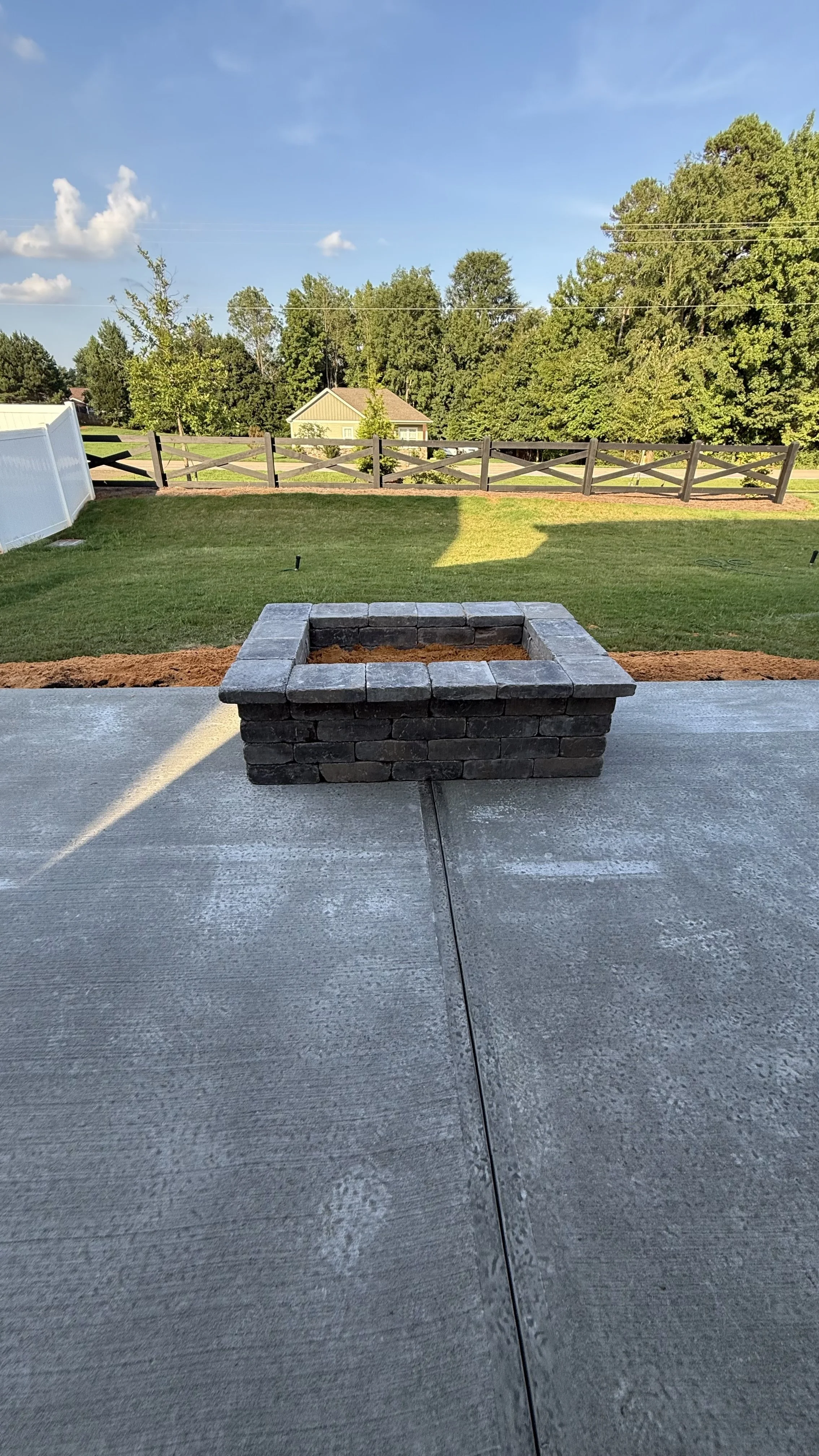 A brick fire pit on a concrete patio in a backyard with green grass, trees, a wooden fence, and a house in the background under a blue sky with some clouds.