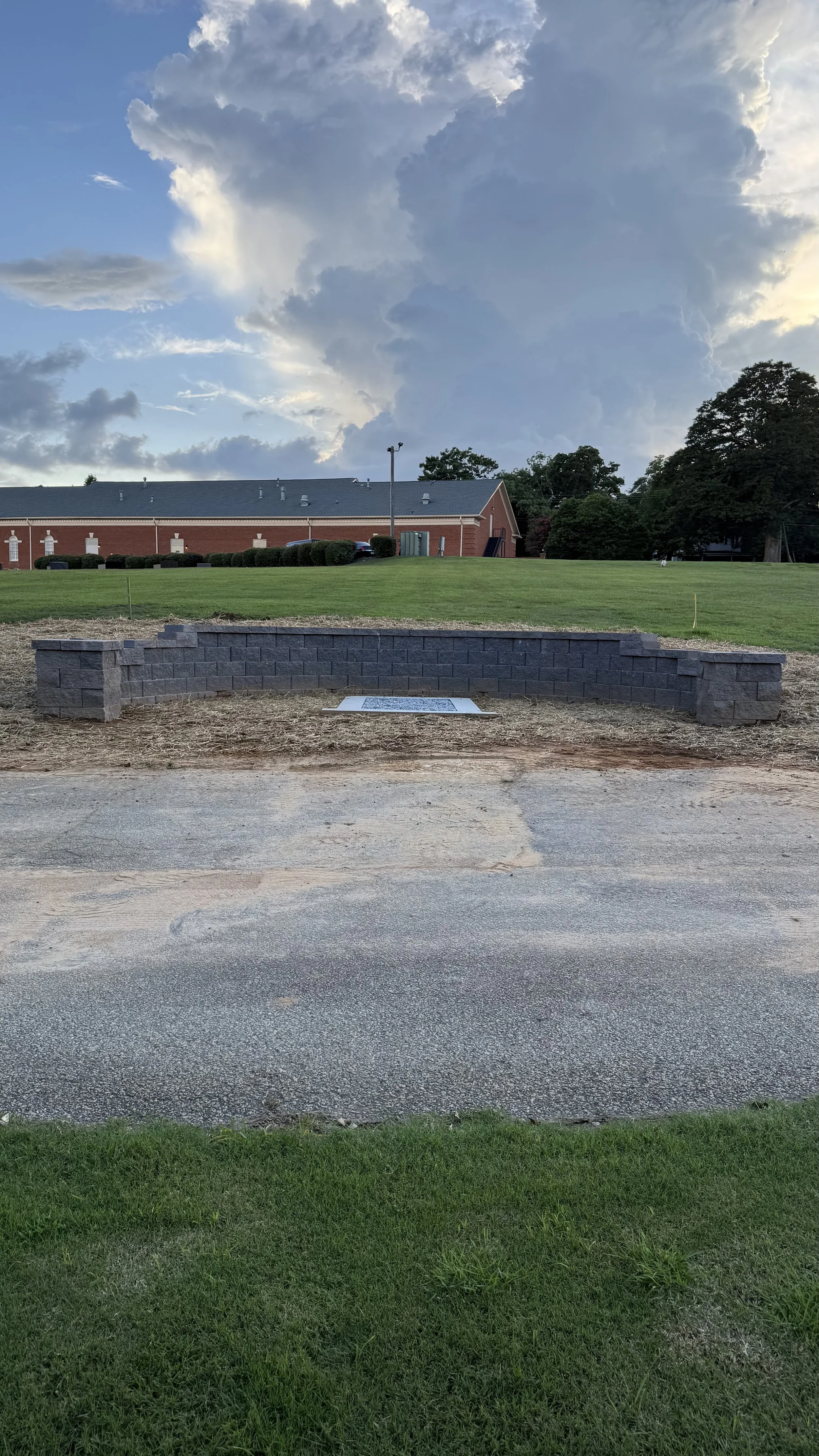A low brick wall with a plaque in front, situated on a grassy area with a large green lawn and a building with a gray roof in the background, under a sky with white and gray clouds.