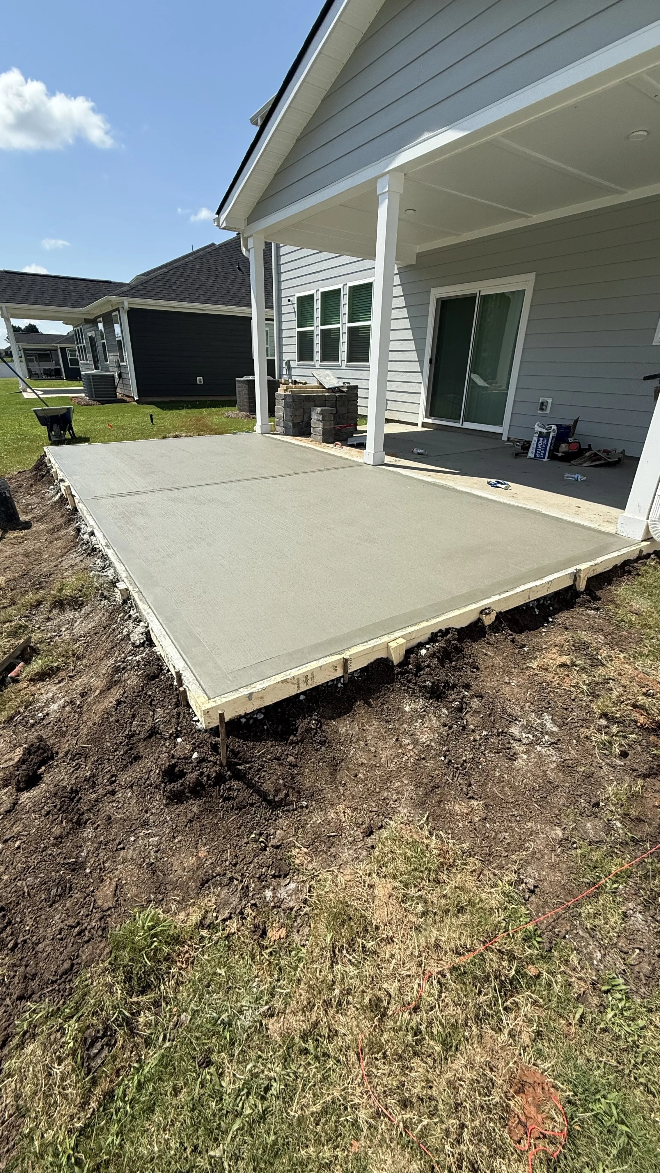 Newly poured concrete patio in the backyard of a suburban house, with construction tools and materials around, grass and neighboring houses in the background, blue sky with a few clouds.