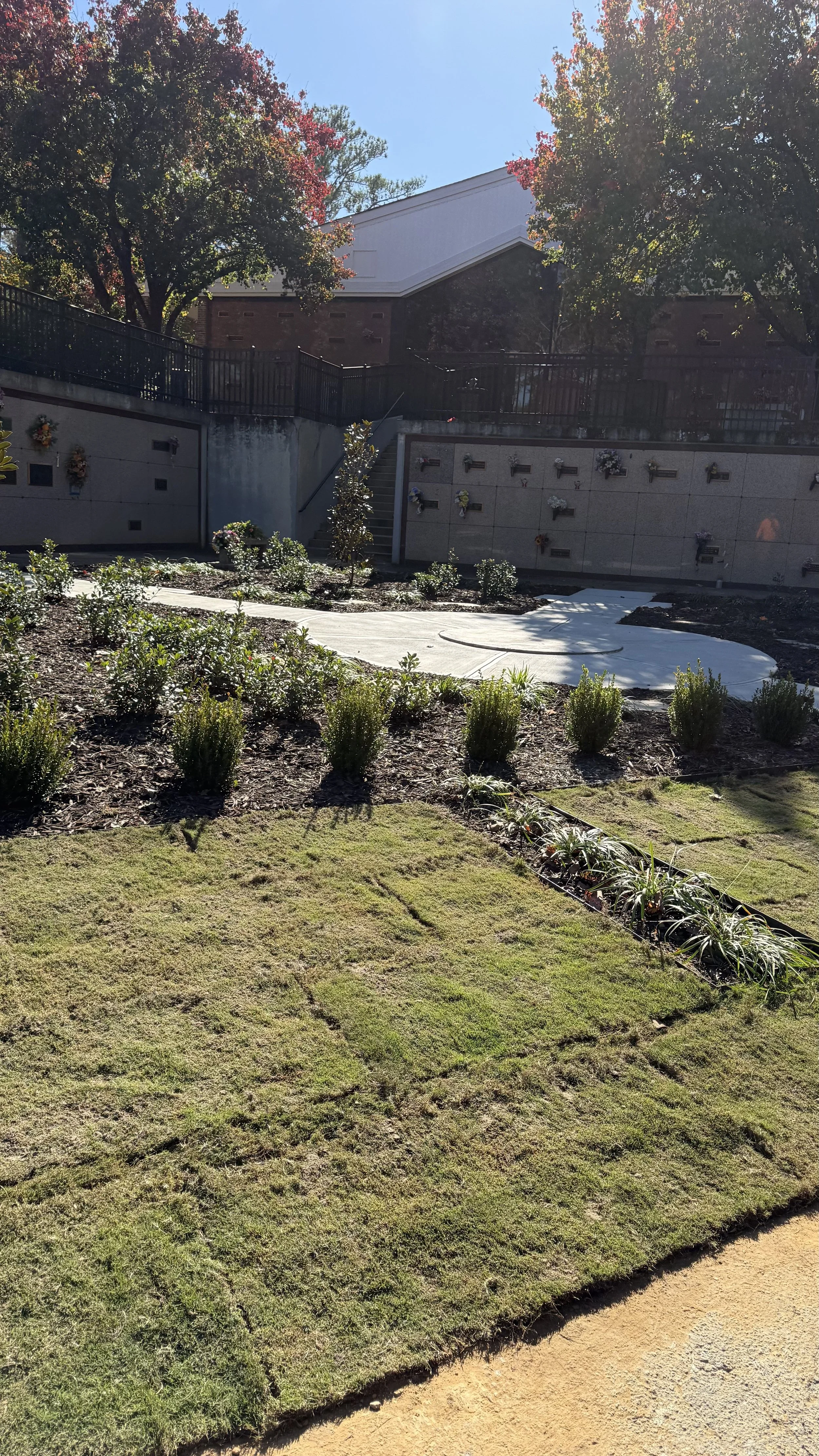 A memorial or gravesite area with a wall featuring multiple plaques and flowers, surrounded by landscaped plants and trees, under a clear blue sky.