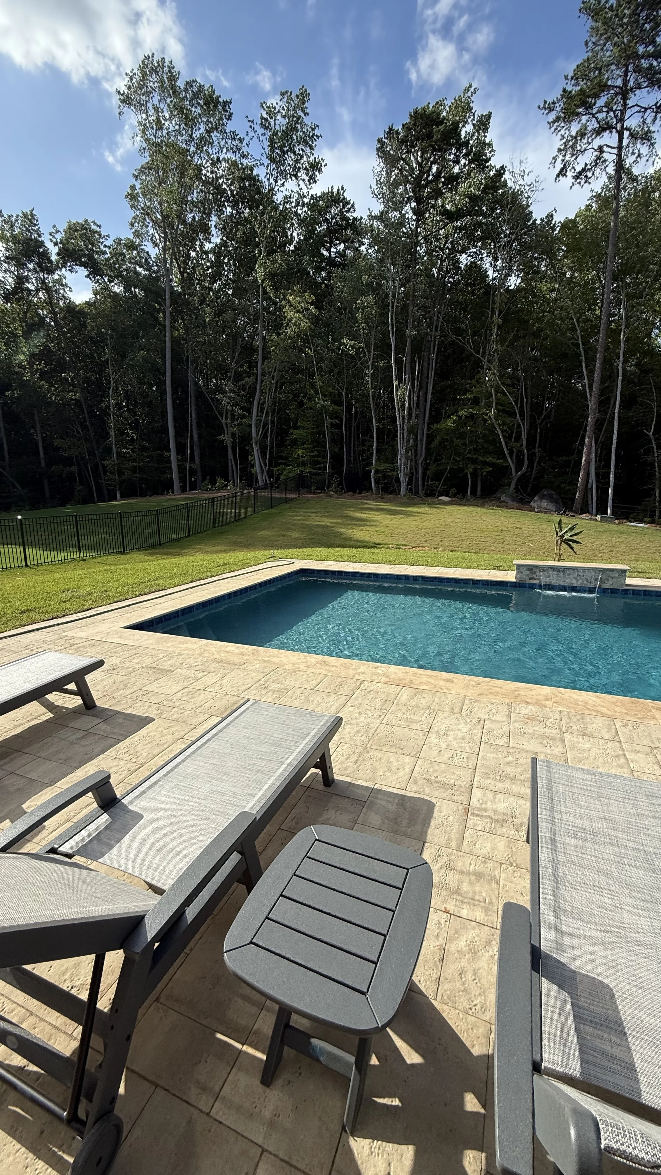 A backyard with a swimming pool, surrounded by a patio with lounge chairs, a grassy area, a black fence, and a background of trees under a partly cloudy sky.