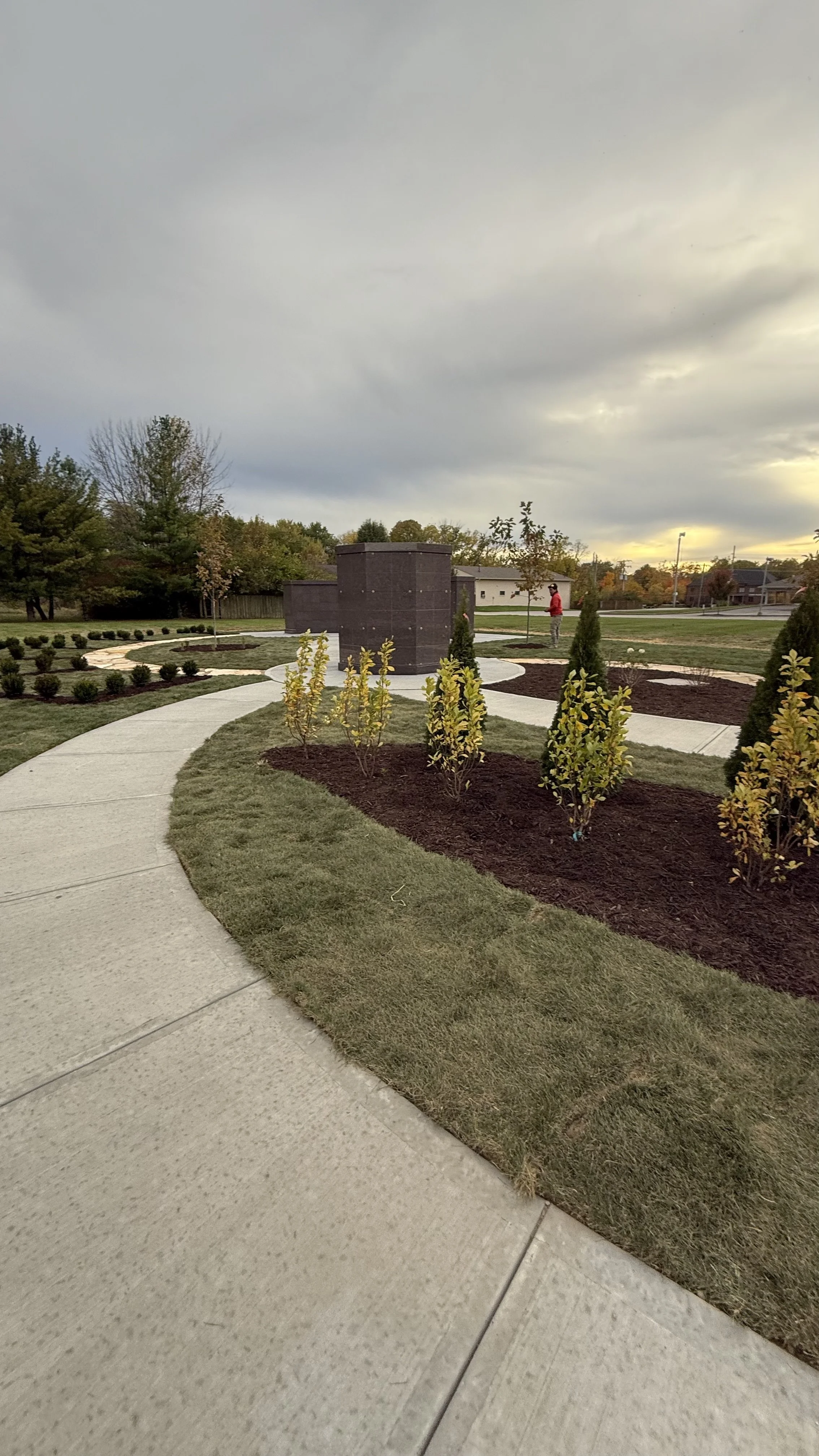 A landscaped park area with a curved concrete pathway, small trees, shrubs, and a monument or structure in the center. There are a few people visible in the background and houses beyond the park, under a cloudy sky.