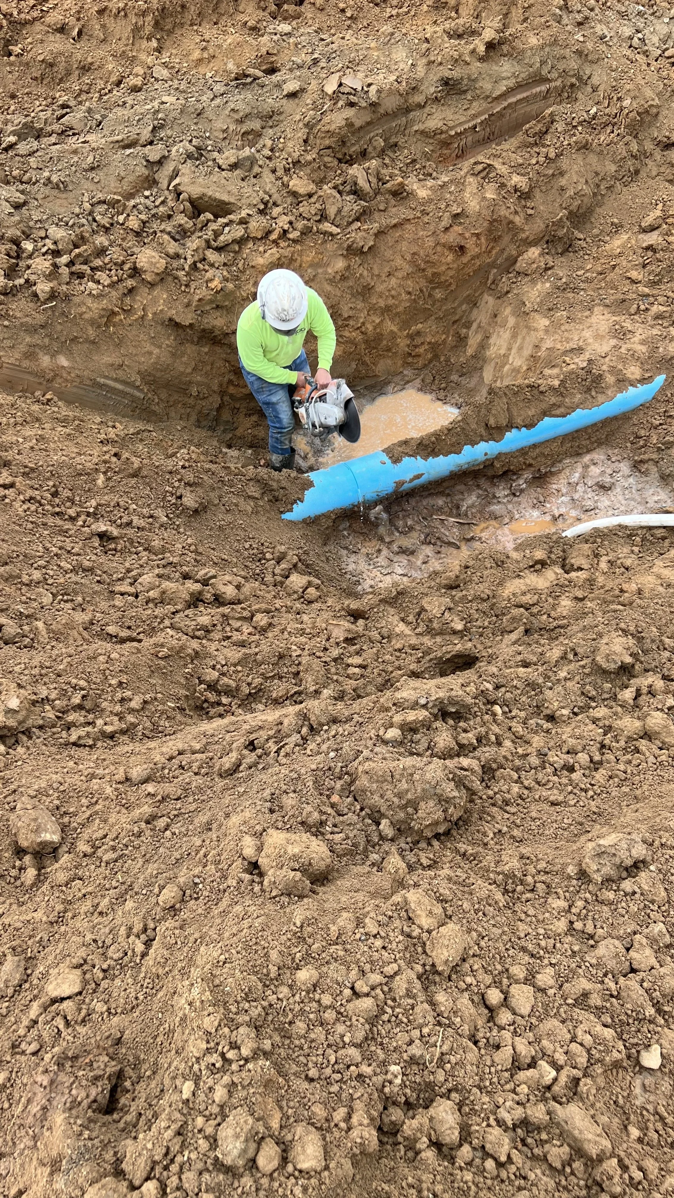 A construction worker with a white helmet and neon green shirt is using a cut-off saw to work on a large blue pipes in a dirt trench.