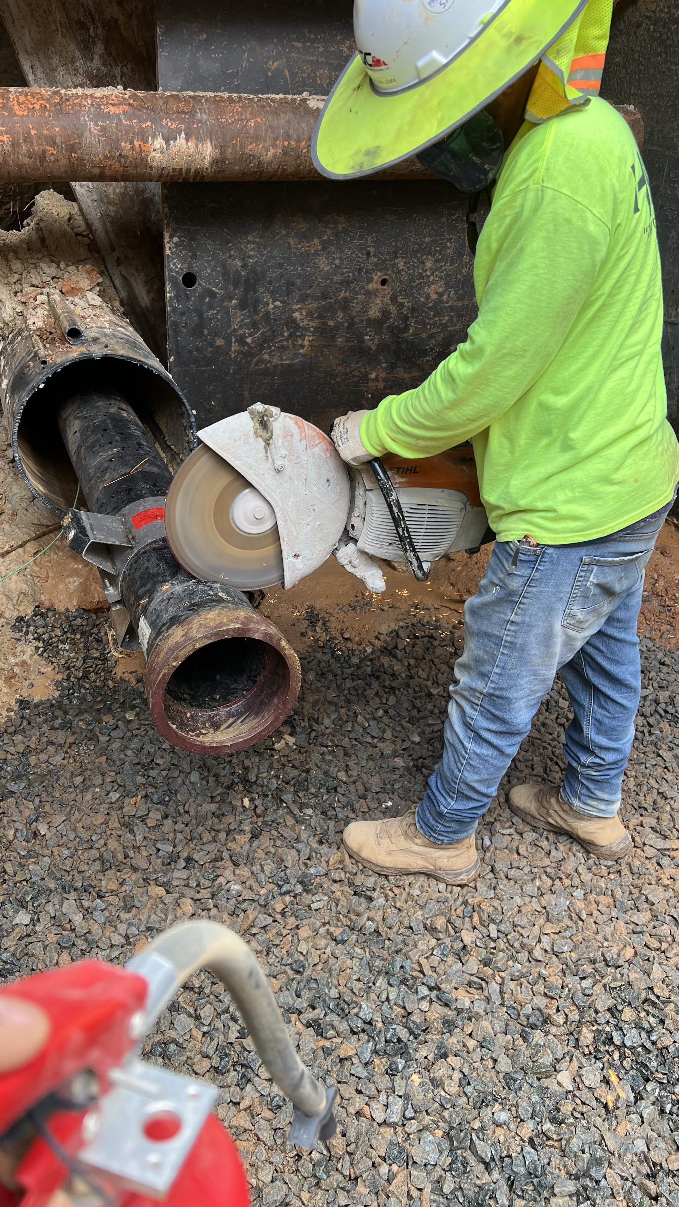 A construction worker in a neon yellow long-sleeve shirt, blue jeans, and safety helmet cuts a large pipe with a circular saw at a construction site with gravel ground and rusted pipes.