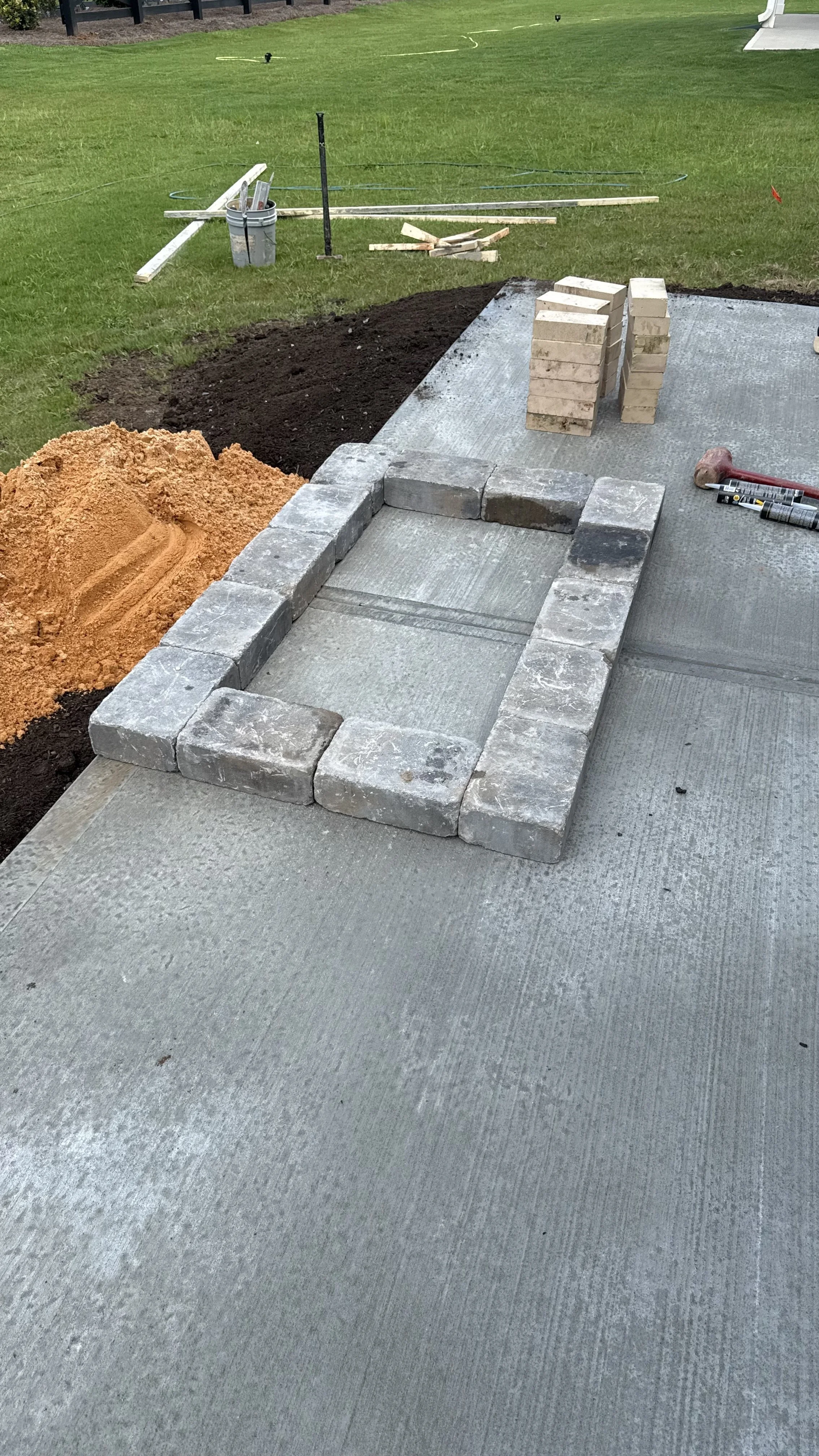 Construction site with stacked bricks, partially built brick structure, and construction tools on concrete patio surrounded by grass.