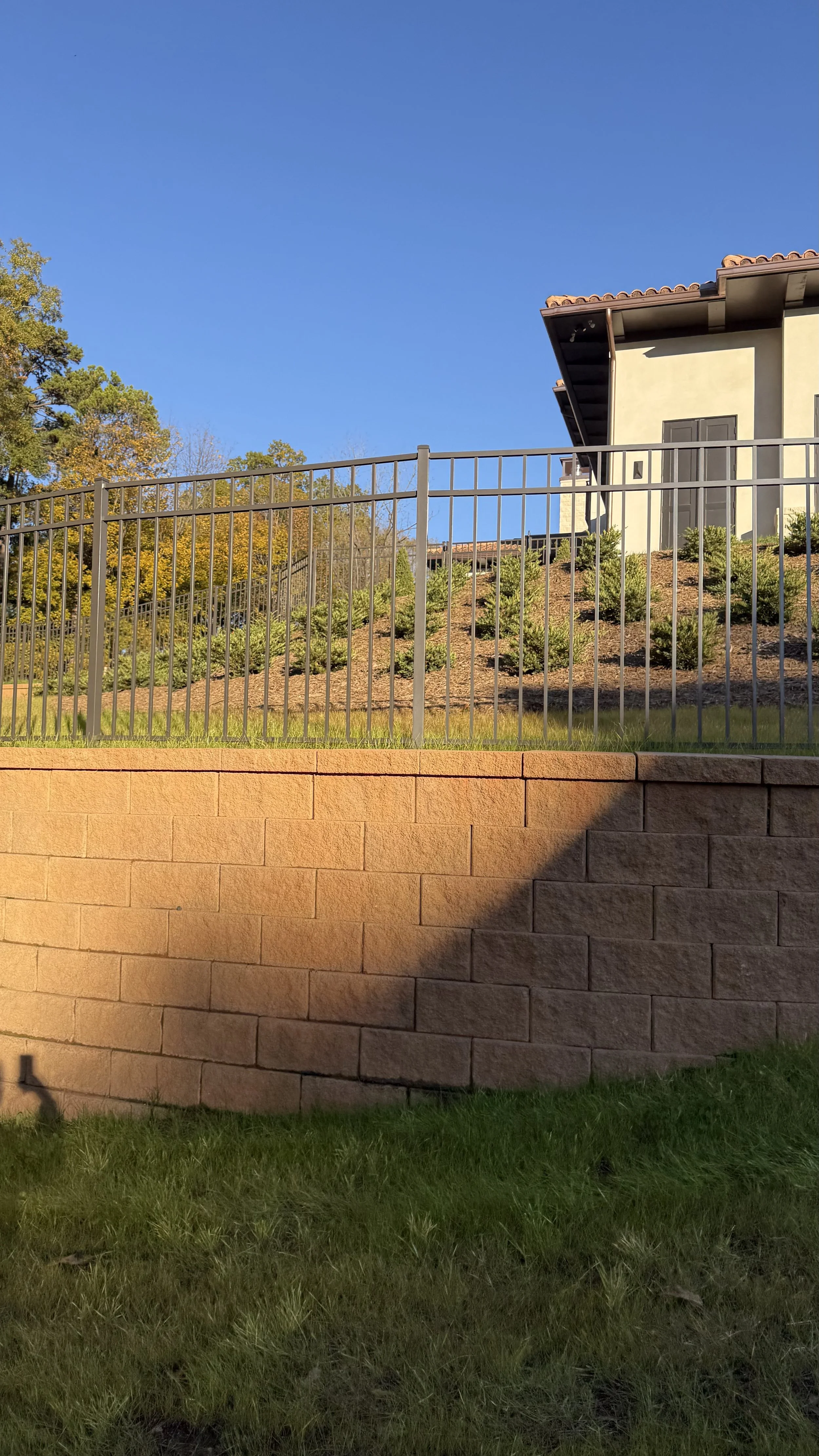 The image shows a house on a hillside with a metal fence, a stone wall in the foreground, and trees with autumn foliage under a clear blue sky.