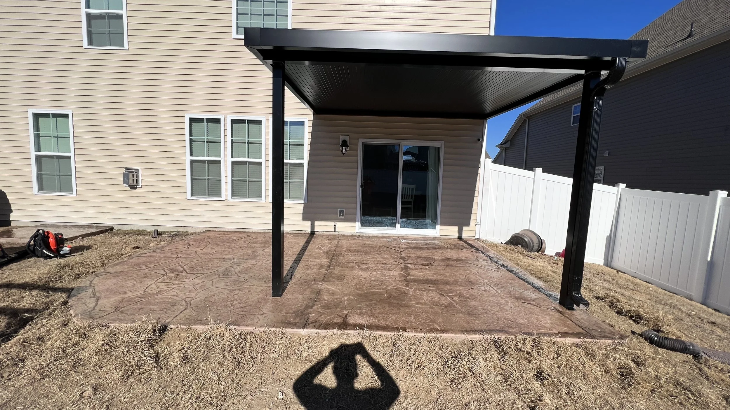 Backyard patio with stamped concrete surface and a covered area with black metal posts, beige house with multiple windows, white fence, and a sliding glass door leading into the house.
