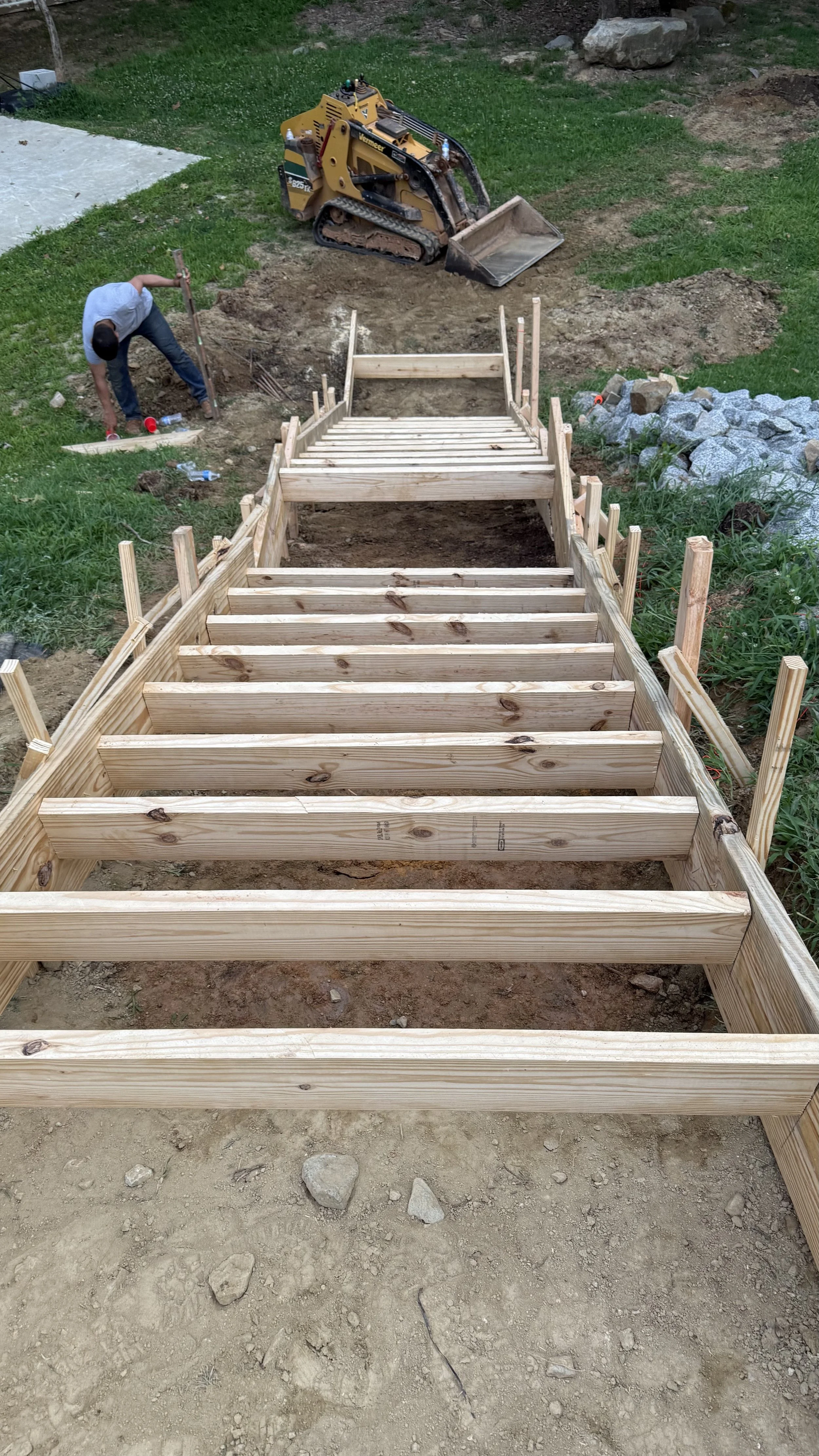 Construction site with wooden stairs framing and a small excavator in the background, working on landscaping or foundation preparation.