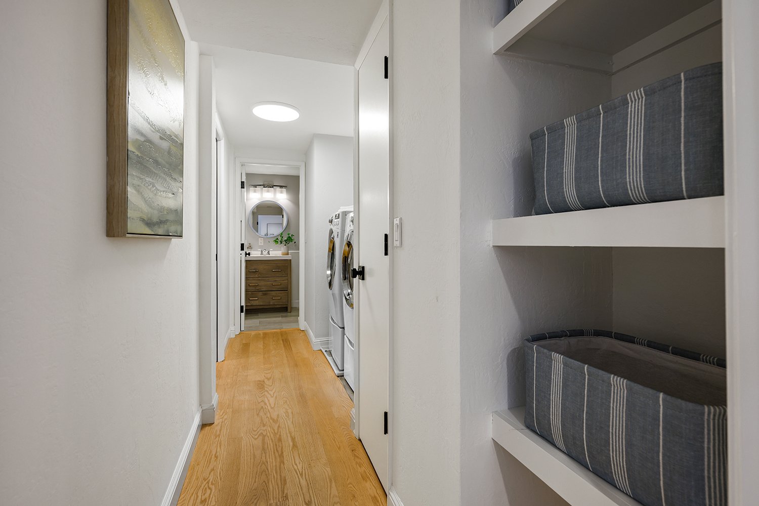 A narrow laundry room with white walls and light wood flooring, featuring a side shelf with gray fabric bins on the right, a white door to the laundry area on the left, and a washer and dryer at the end of the room. There is a small vanity with a mir