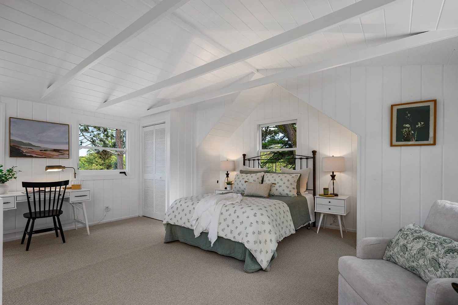 Bright bedroom with white paneled walls and ceiling, featuring a bed with white floral bedding, two nightstands with lamps, a white dresser with a desk and chair, window views of trees, and a framed artwork on the wall.