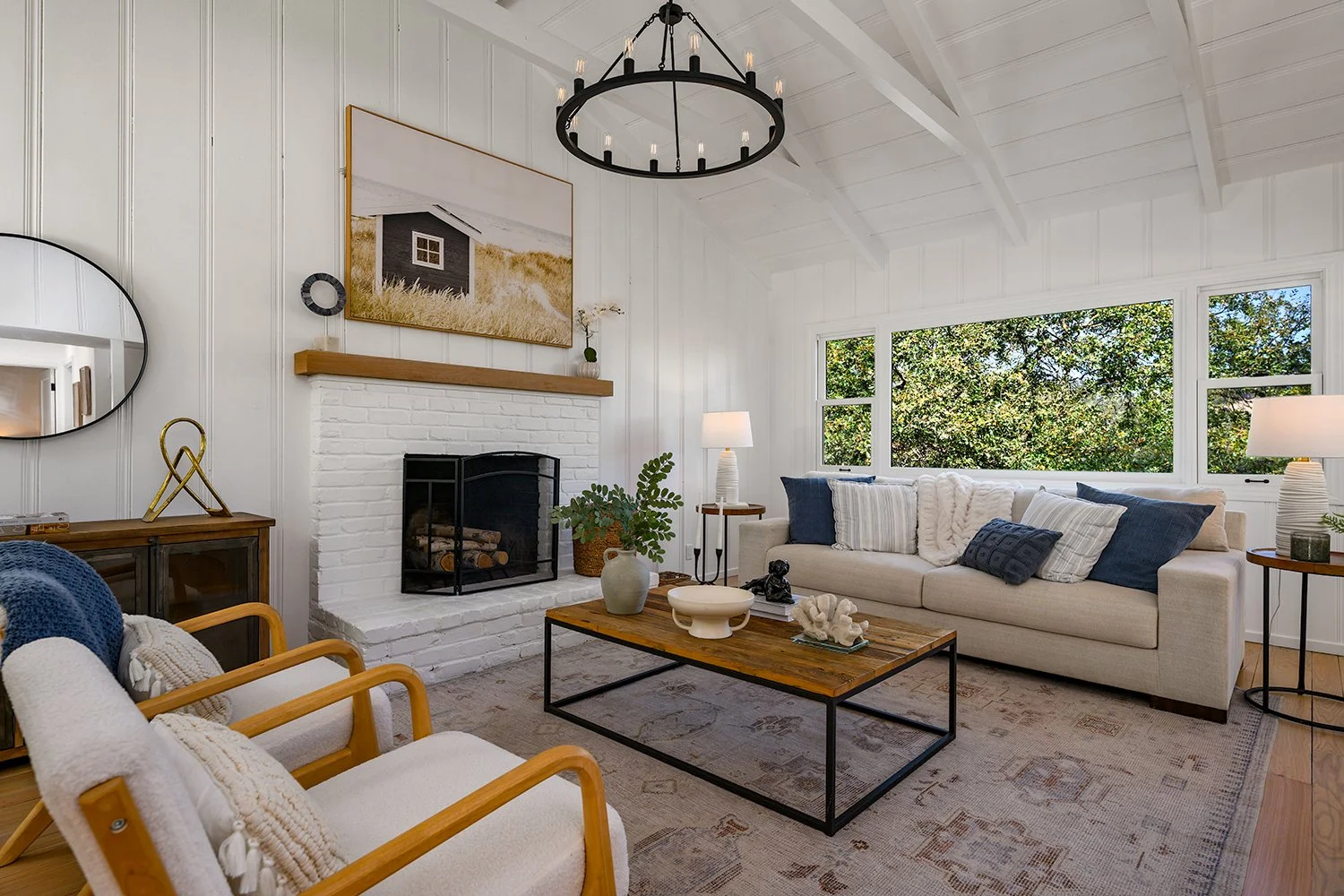 Bright living room with white walls and ceiling, large windows showing green trees outside, a white sofa with blue and white pillows, wooden armchairs with beige cushions, a rustic coffee table with decorative items, a black and white framed photogra