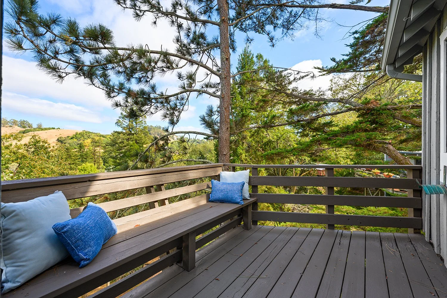 A wooden balcony with a bench and pillows, overlooking trees and hills in the distance under a partly cloudy sky.