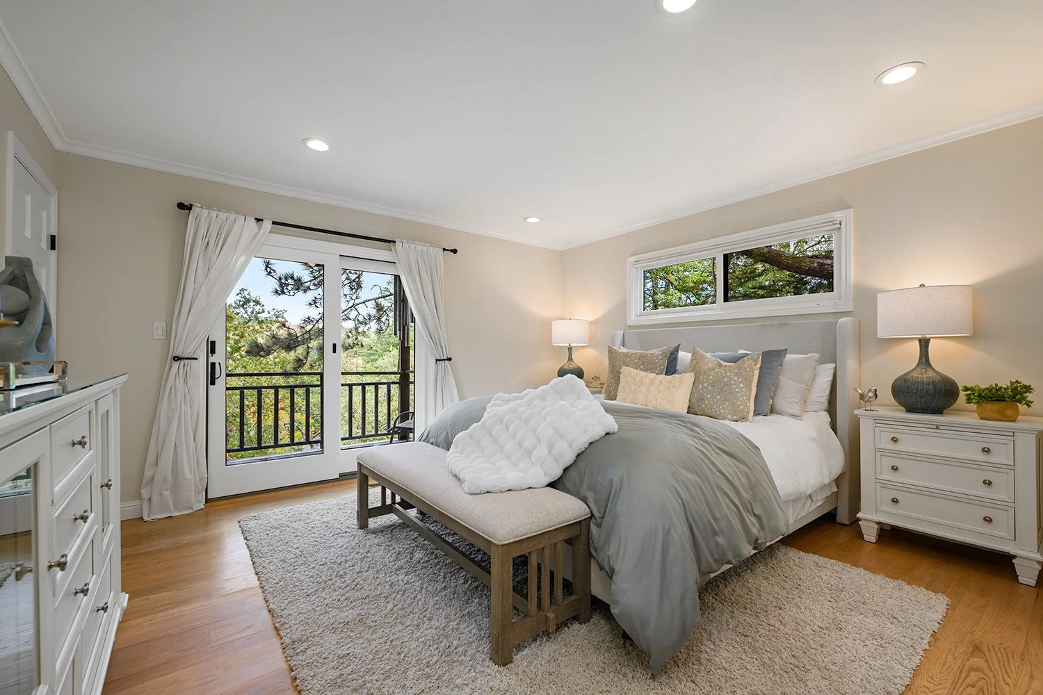 A cozy bedroom with a large bed, white and beige pillows, and a gray comforter. There's a bench at the foot of the bed, a white nightstand with a lamp and decorative items, and sliding glass doors leading to a balcony with trees outside. A window ove