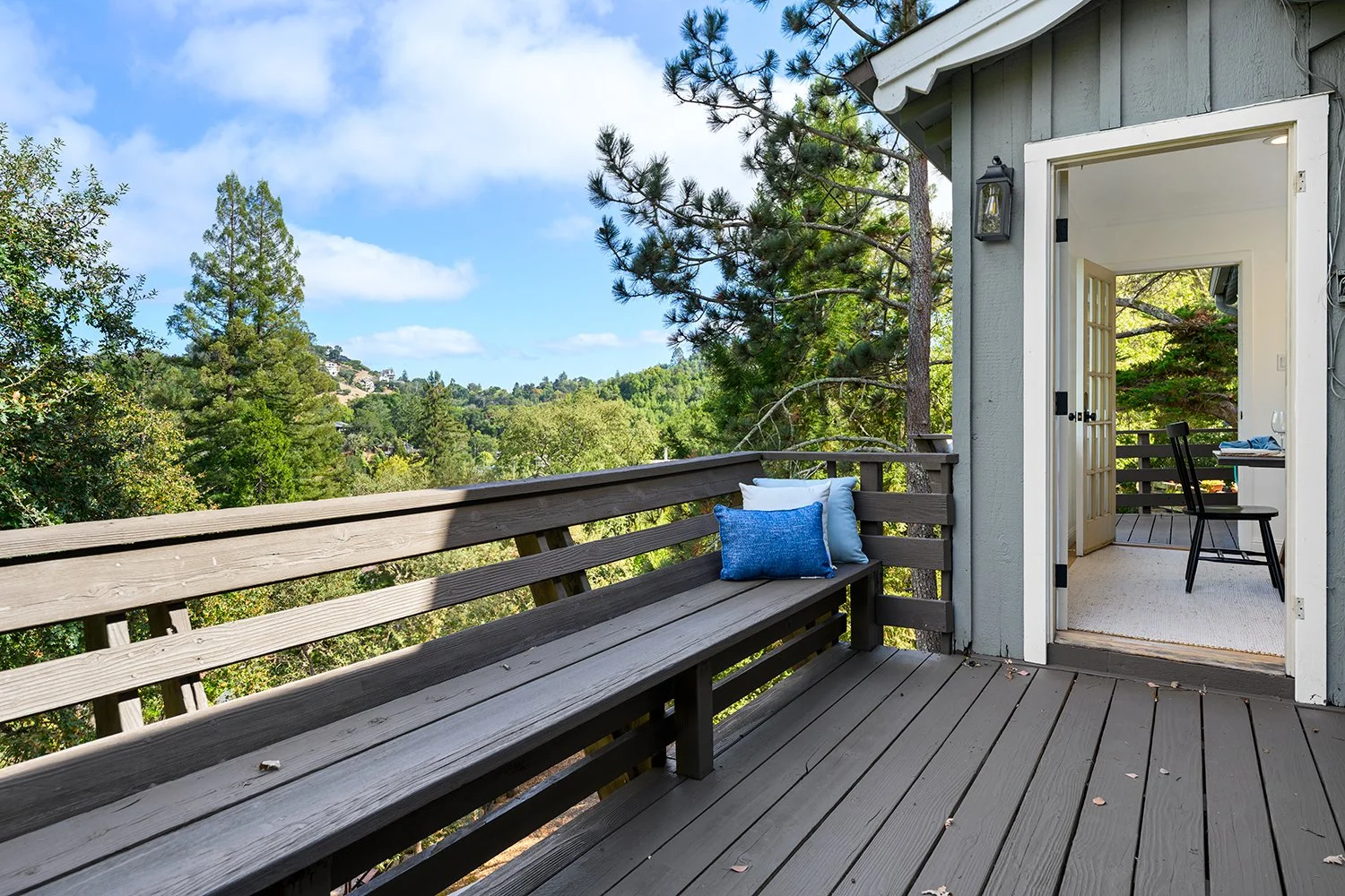 A wooden outdoor balcony with a bench and blue pillows, overlooking green trees and a hilly landscape, connected to a house with an open door leading inside.