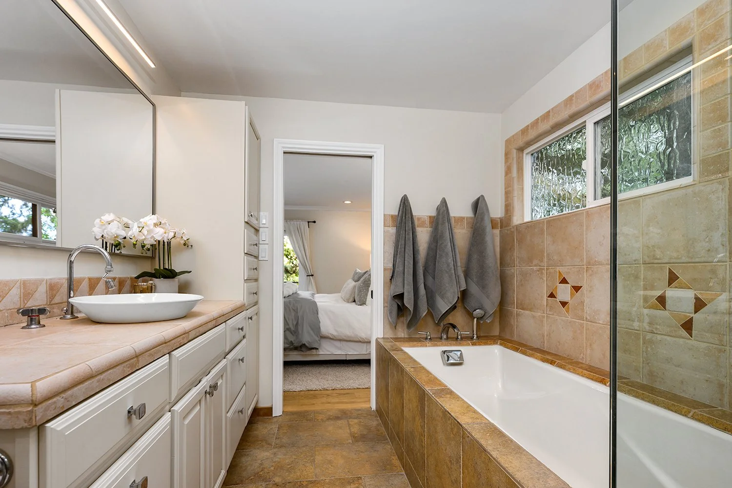 Bathroom with beige tiled bathtub and wall, window, and white vanity with sink, mirror, orchid plant, and towels