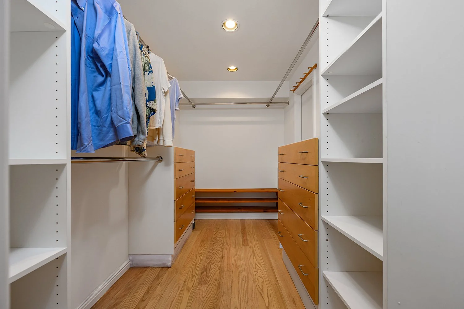 Empty walk-in closet with wooden drawers and built-in shelves, with hanging clothes on the left side and tracks for additional hanging rods.