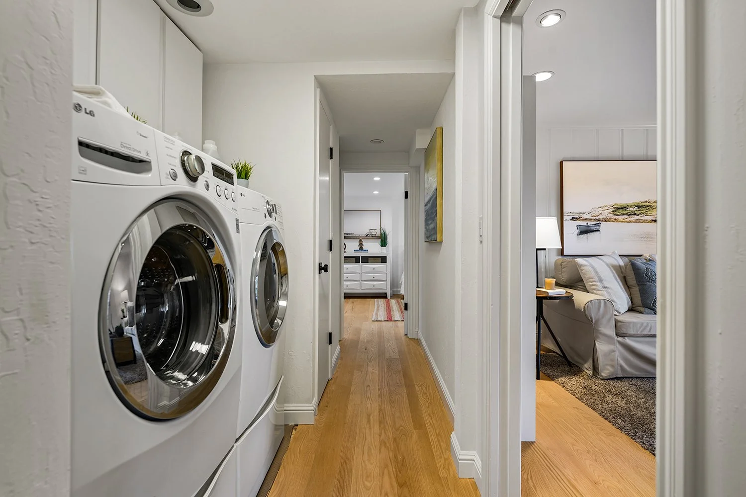 View down a hallway with laundry room on the left containing stacked washer and dryer, and a living room on the right with a couch, lamp, and large landscape painting on the wall.