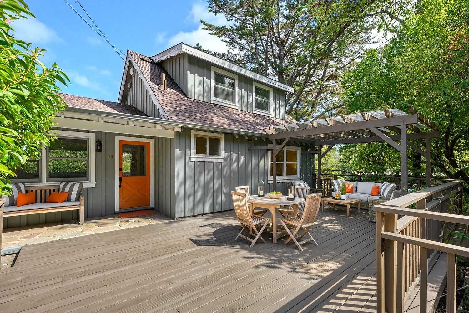 A spacious outdoor deck attached to a house with a gray exterior, orange door, and multiple windows. The deck has outdoor furniture including a round table with four chairs, a corner sofa with striped cushions, and several orange and striped throw pi