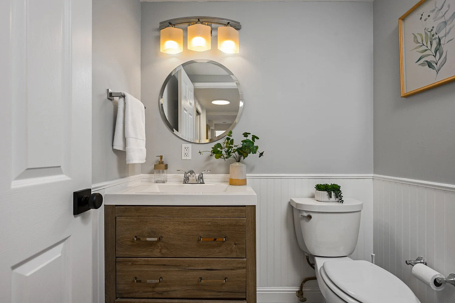 A modern bathroom featuring a white vanity with a wooden cabinet, a round mirror, a three-light fixture, a white toilet with a small plant on top, and a white towel hanging on the wall.