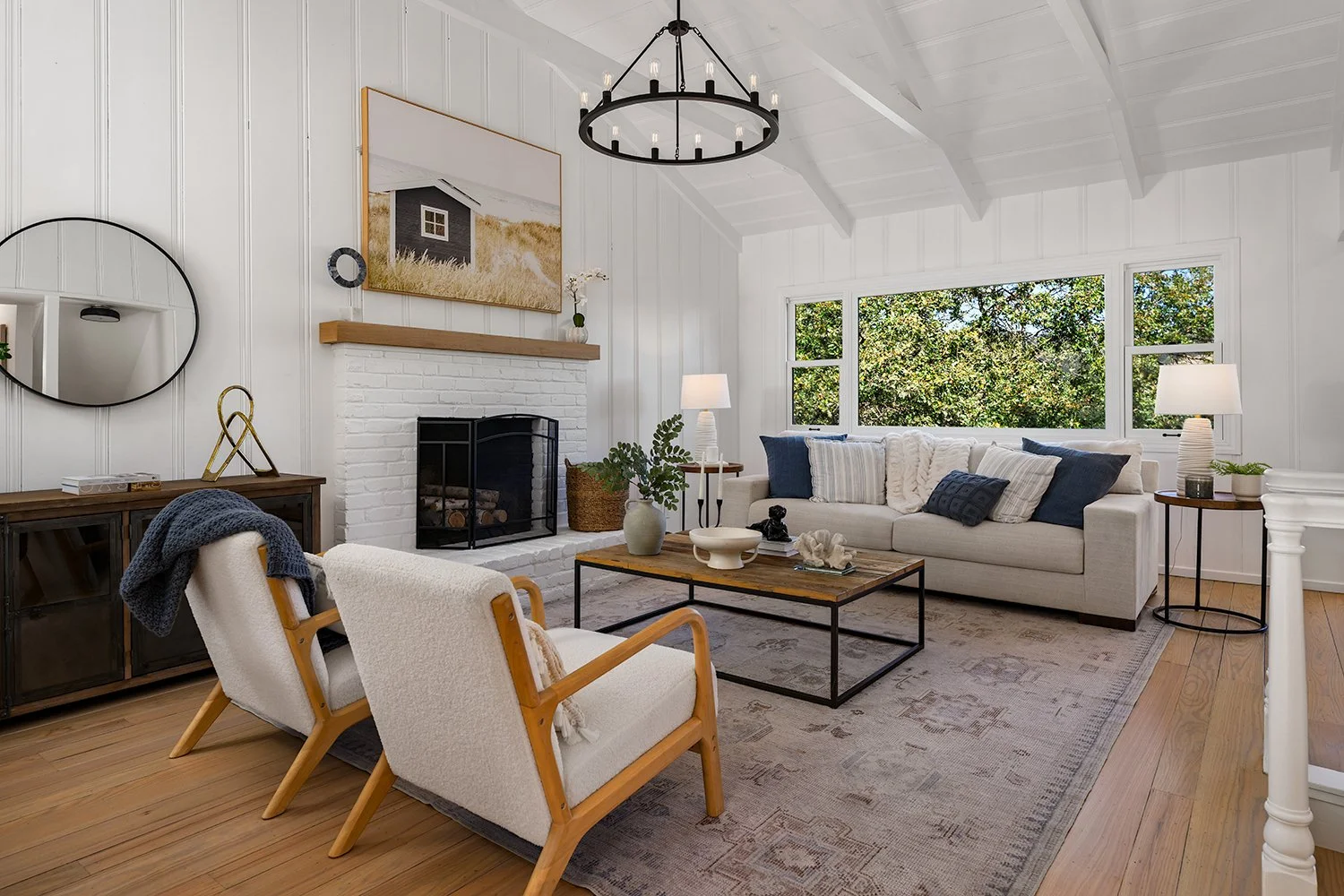 A living room with white walls, a white brick fireplace, a large window with green trees outside, a beige sofa with blue and white pillows, two cream-colored armchairs, a wooden coffee table, and side tables with lamps.