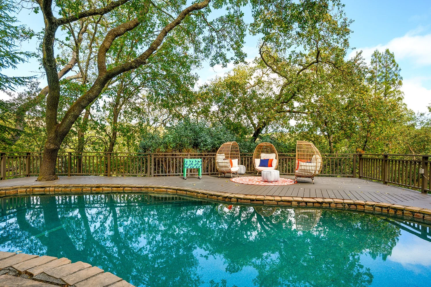 A backyard swimming pool with outdoor furniture, surrounded by trees and greenery, on a clear day.