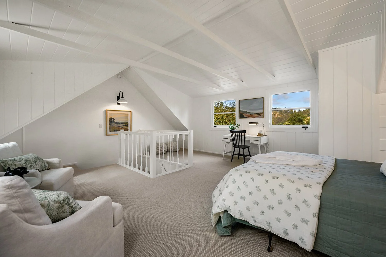 A spacious attic bedroom with white walls and ceiling, featuring a bed with a floral blanket, a white armchair with pillows, a small desk with a black chair, and a view of trees through three windows.