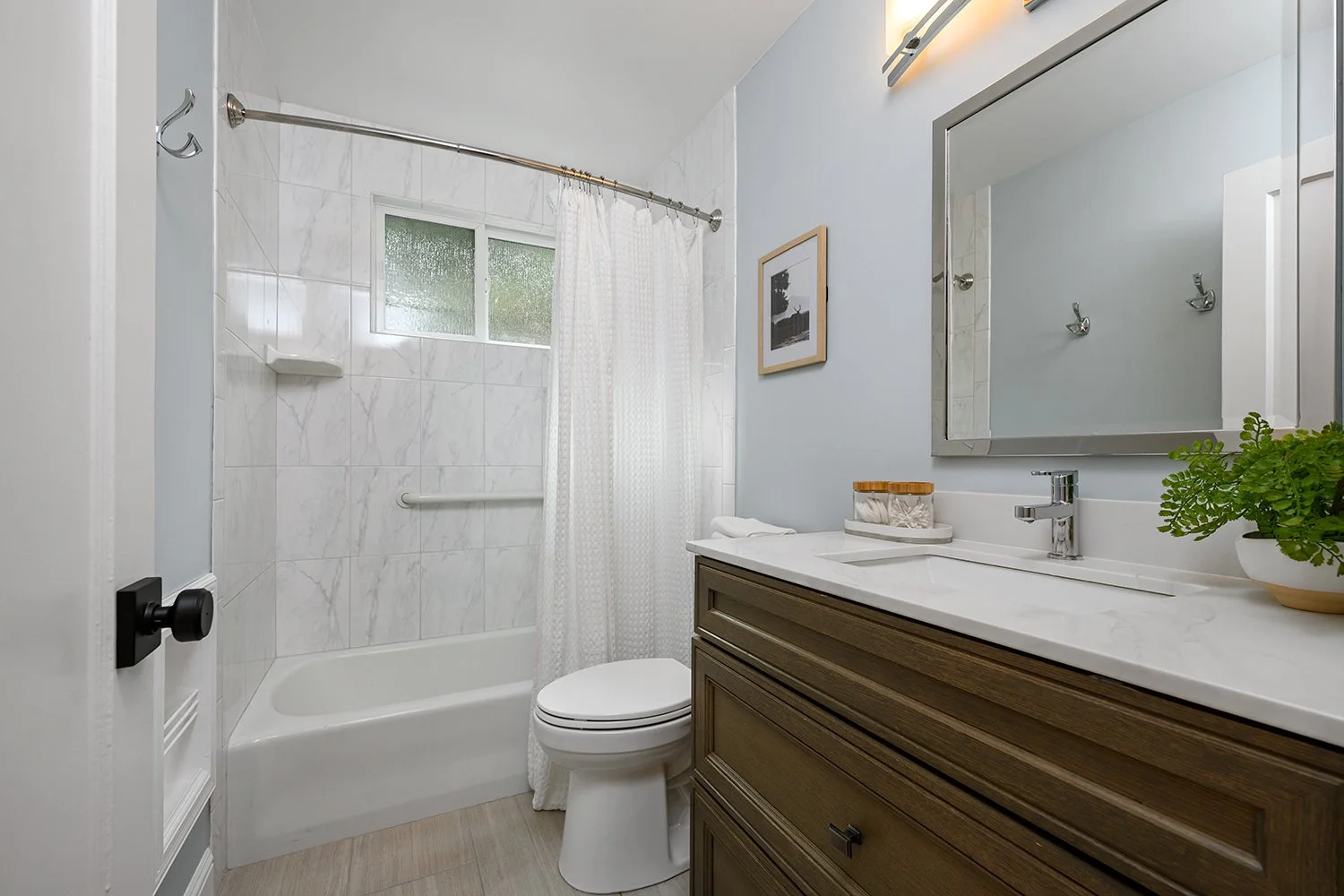 Bathroom with bathtub, white shower curtain, window with frosted glass, toilet, wooden vanity with white countertop, mirror, potted plant, framed picture on light blue wall.