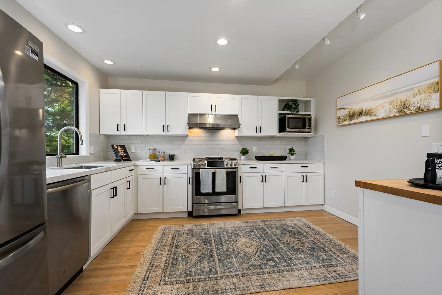 Modern kitchen with white cabinets, stainless steel appliances, a window with green trees outside, and a wooden floor. There is a patterned rug on the floor and decorative plants on the counter.