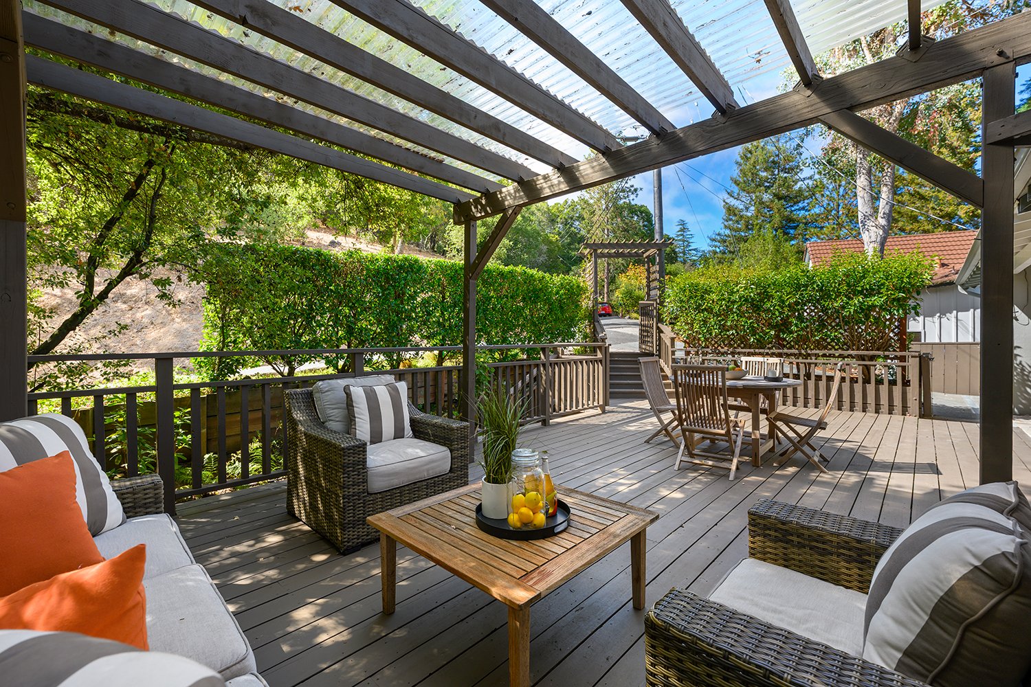 A wooden outdoor patio with a seating area including a cushioned sofa with orange and striped pillows, rattan armchairs, and a wooden dining table with chairs. There is a tray on the coffee table with lemons and glass jars. The patio is shaded by a p