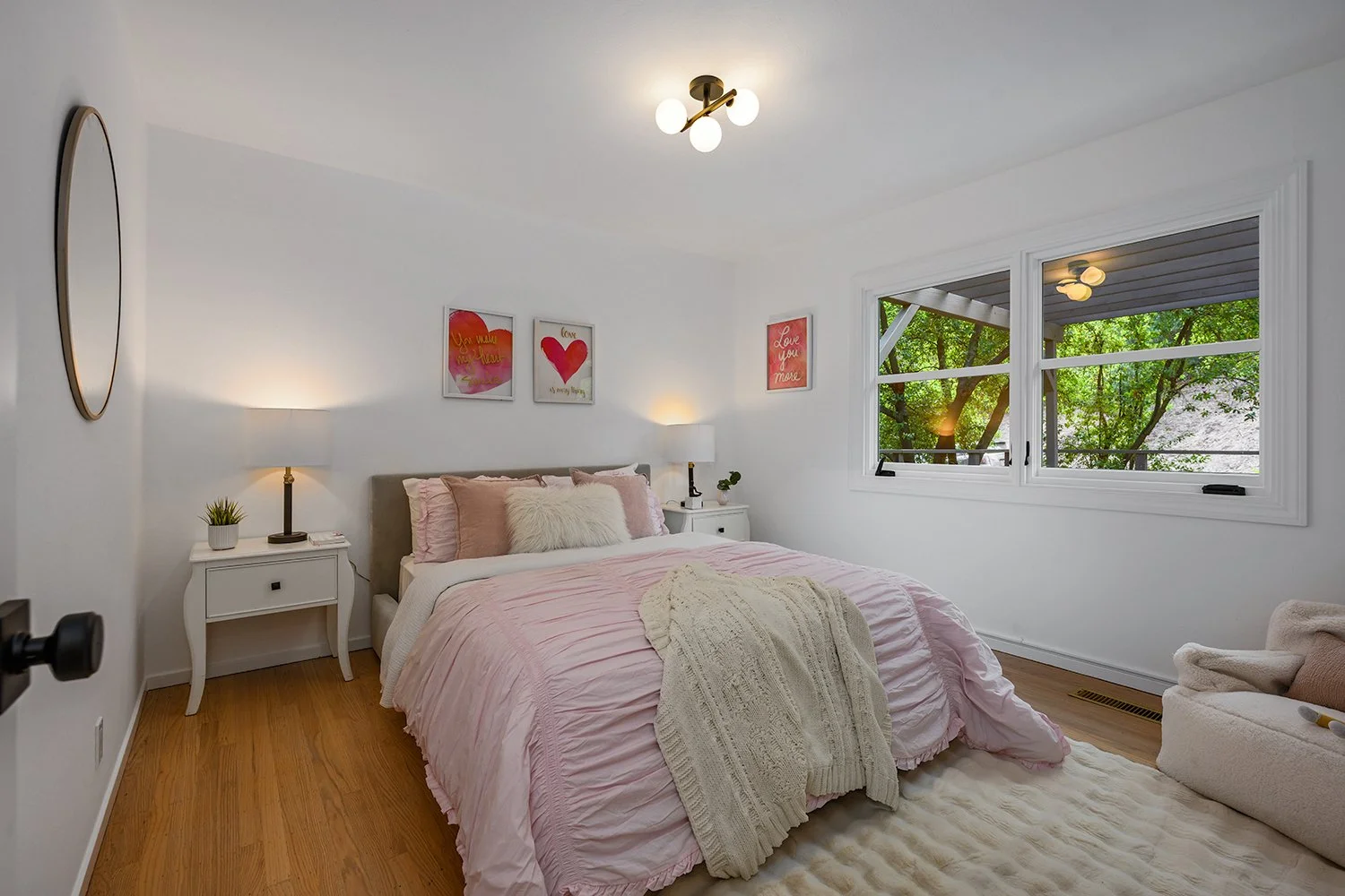 A cozy bedroom with a large bed covered in pink bedding and decorative pillows. There are two white nightstands with lamps on each side of the bed, and a window showing green trees outside. The wall above the bed has three pink and red framed art pie