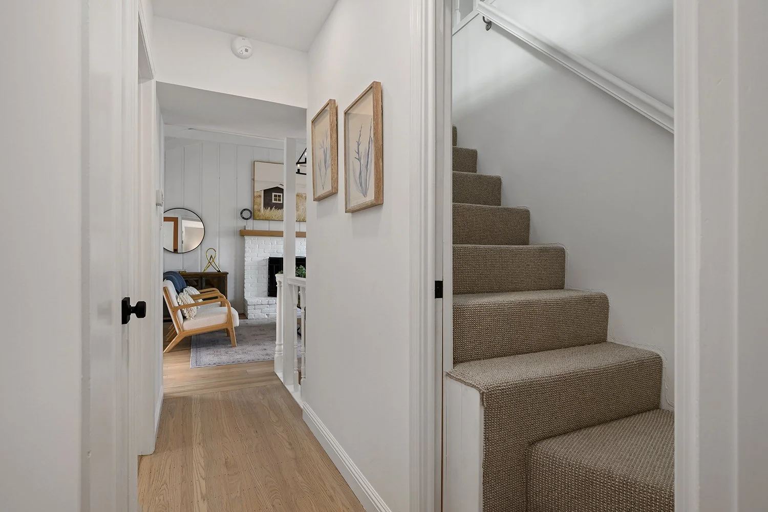 View of a hallway leading into a living room with a white brick fireplace, armchair, and wall art, with a carpeted staircase on the right.