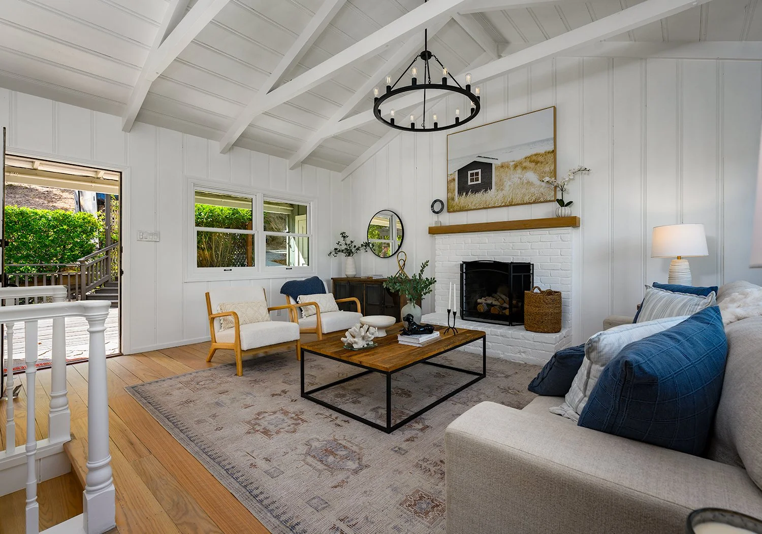 Living room with white walls, vaulted ceiling, black chandelier, brick fireplace, beige sofa with blue pillows, wooden armchairs, coffee table, and large window showing greenery outside.