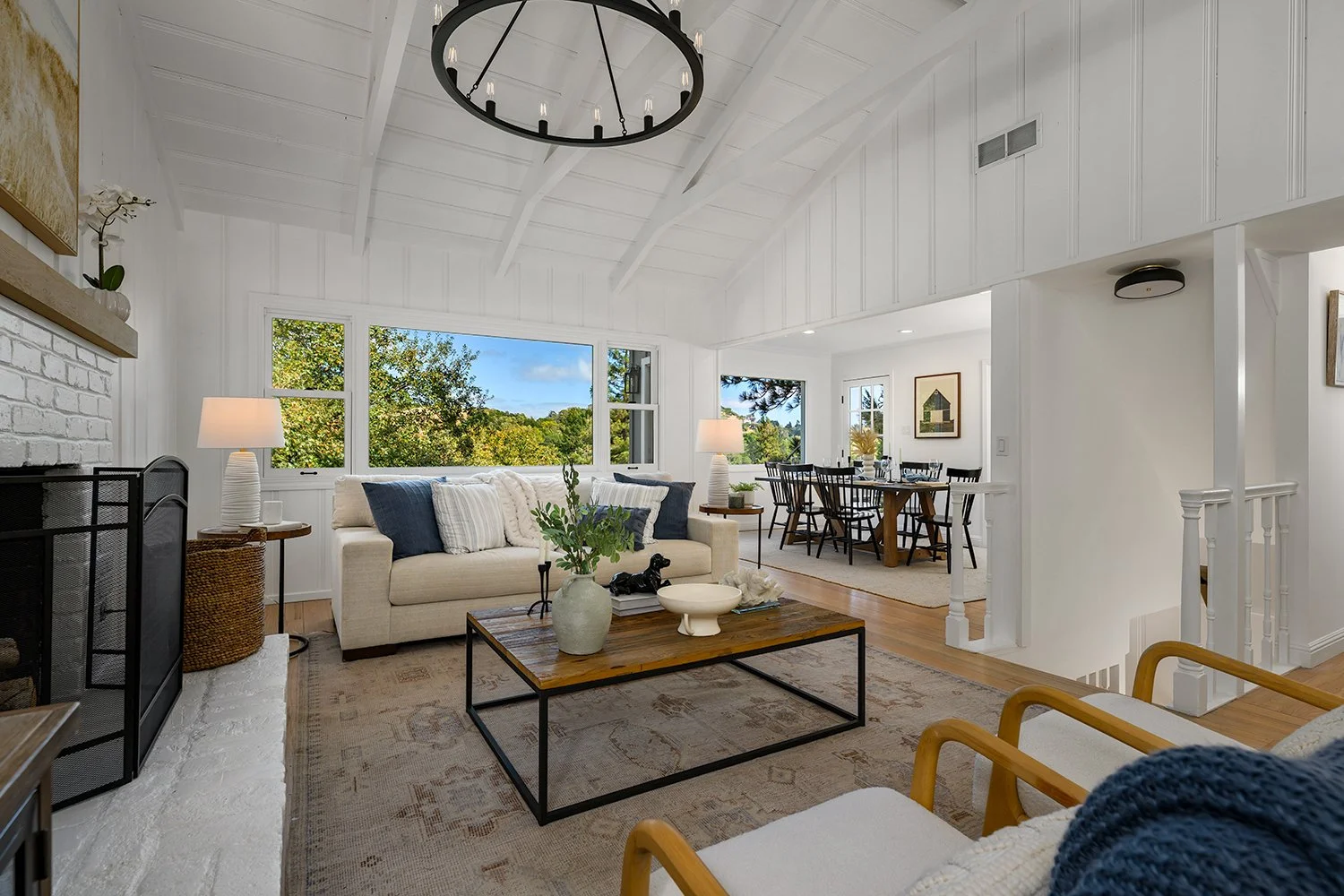 Bright living room with white walls and vaulted ceiling, large window showing green trees, beige sofa with blue and white pillows, wooden coffee table with decorative items, and a dining area with a table and black chairs in the background.