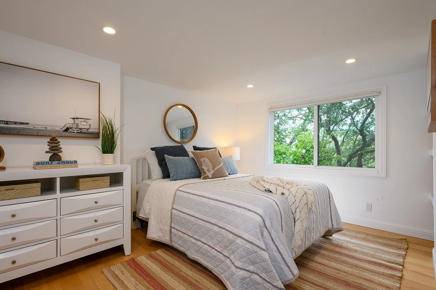 Bright bedroom with white walls, a large window showing green trees, a white bed with striped bedding, multiple pillows, a wooden dresser with decor, a round mirror, and a striped rug.