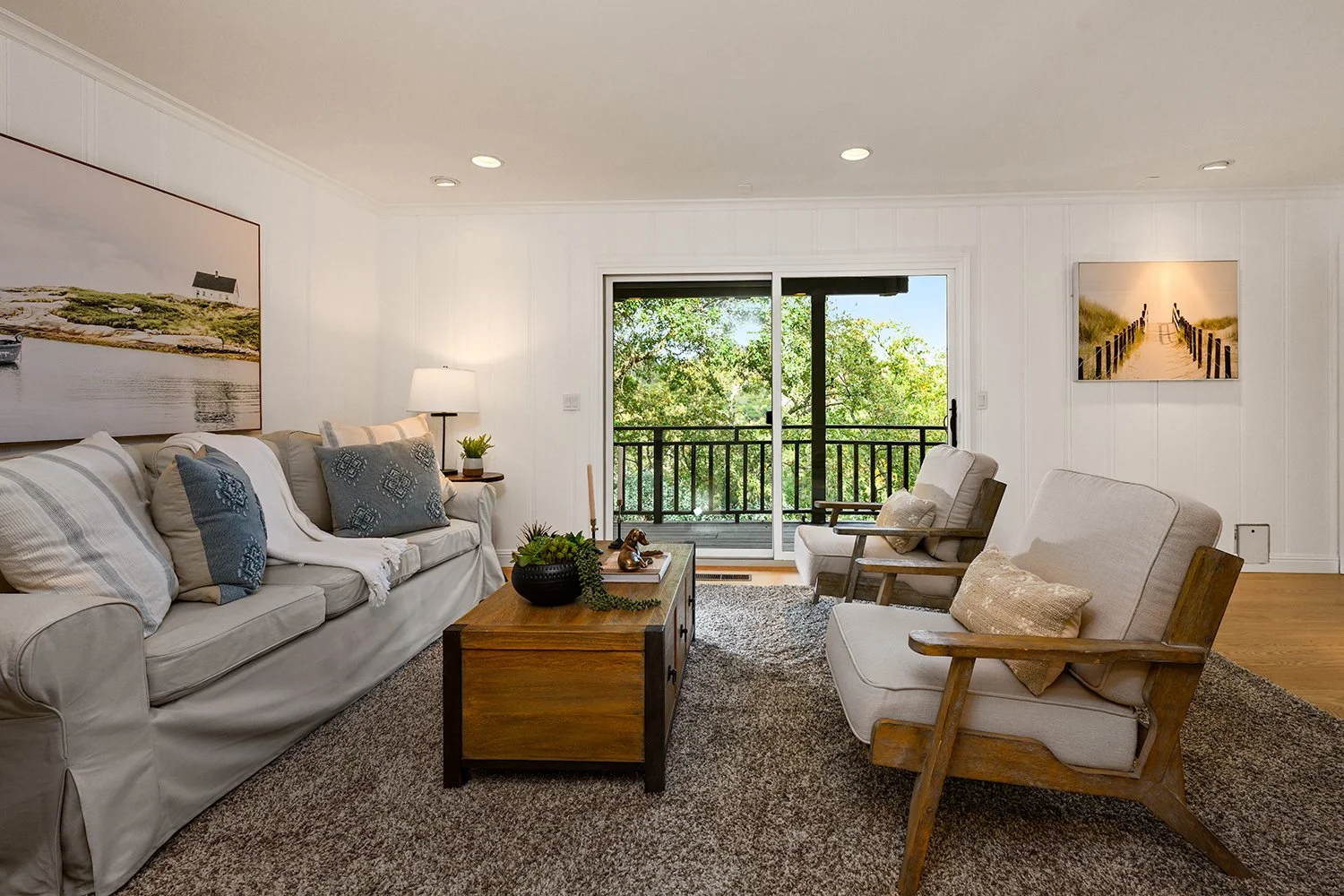 Living room with beige sofa, wooden chairs, a coffee table, and a sliding glass door opening to a balcony with greenery.
