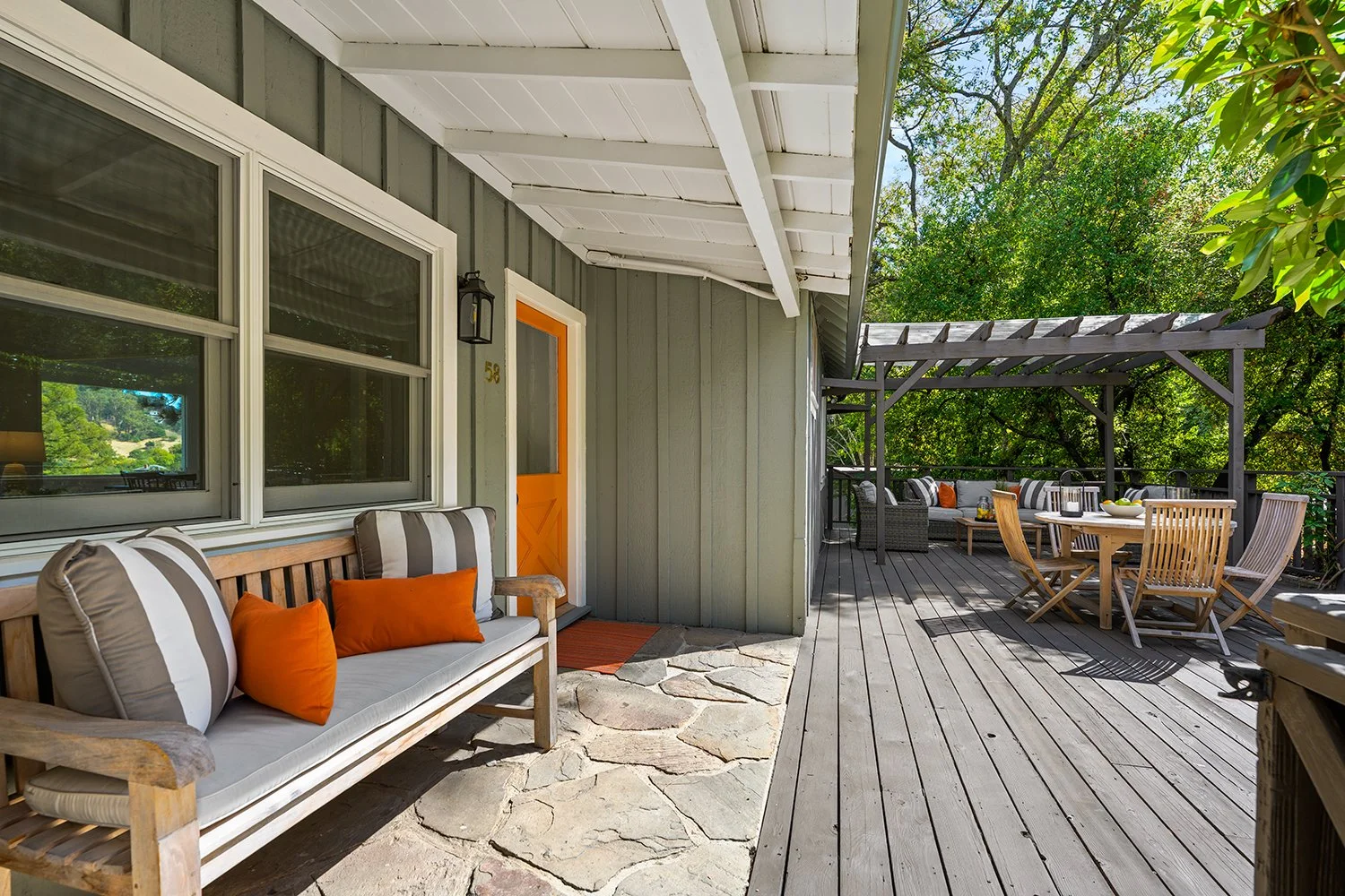 A spacious outdoor wooden deck with a green house wall, a sitting area with a wooden bench and orange pillows, a dining table with chairs, and lush trees in the background.