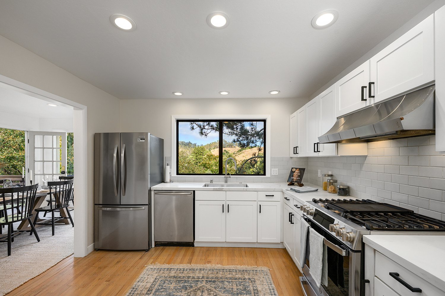 Modern kitchen with white cabinets, stainless steel appliances, a window with a view of trees and hills, and hardwood floors.